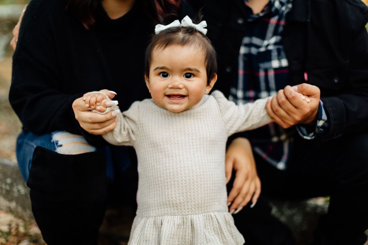 Toddler in cream sweater dress and white bow smiles while being held by adults outdoors.
