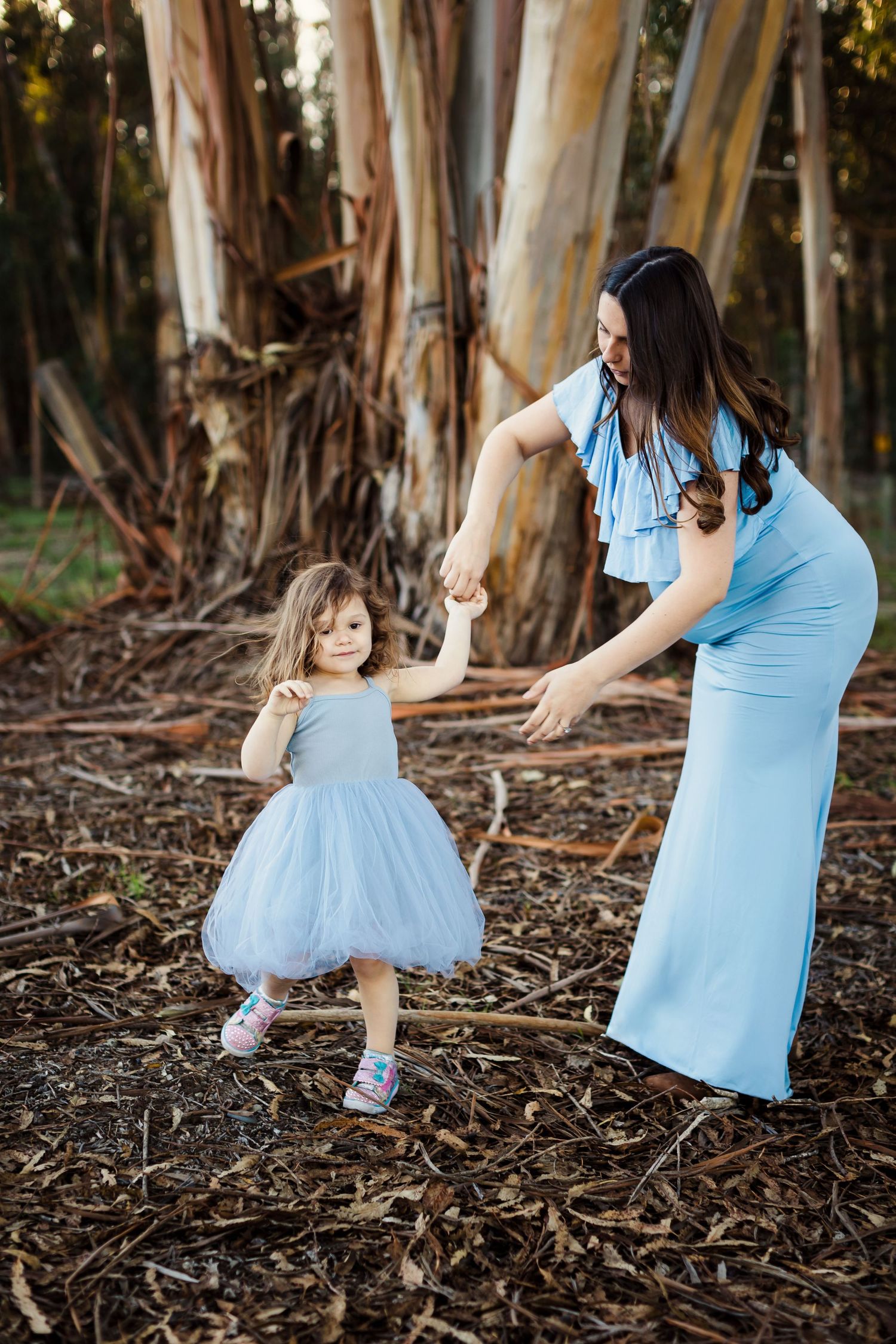 A mother and daughter in matching light blue dresses dance together on fallen eucalyptus leaves in a forest.