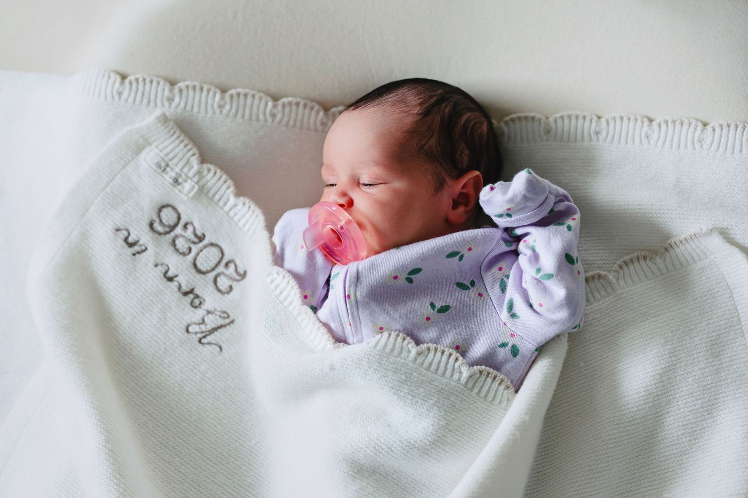 Newborn baby sleeping peacefully wrapped in a white knit blanket with a pink pacifier.