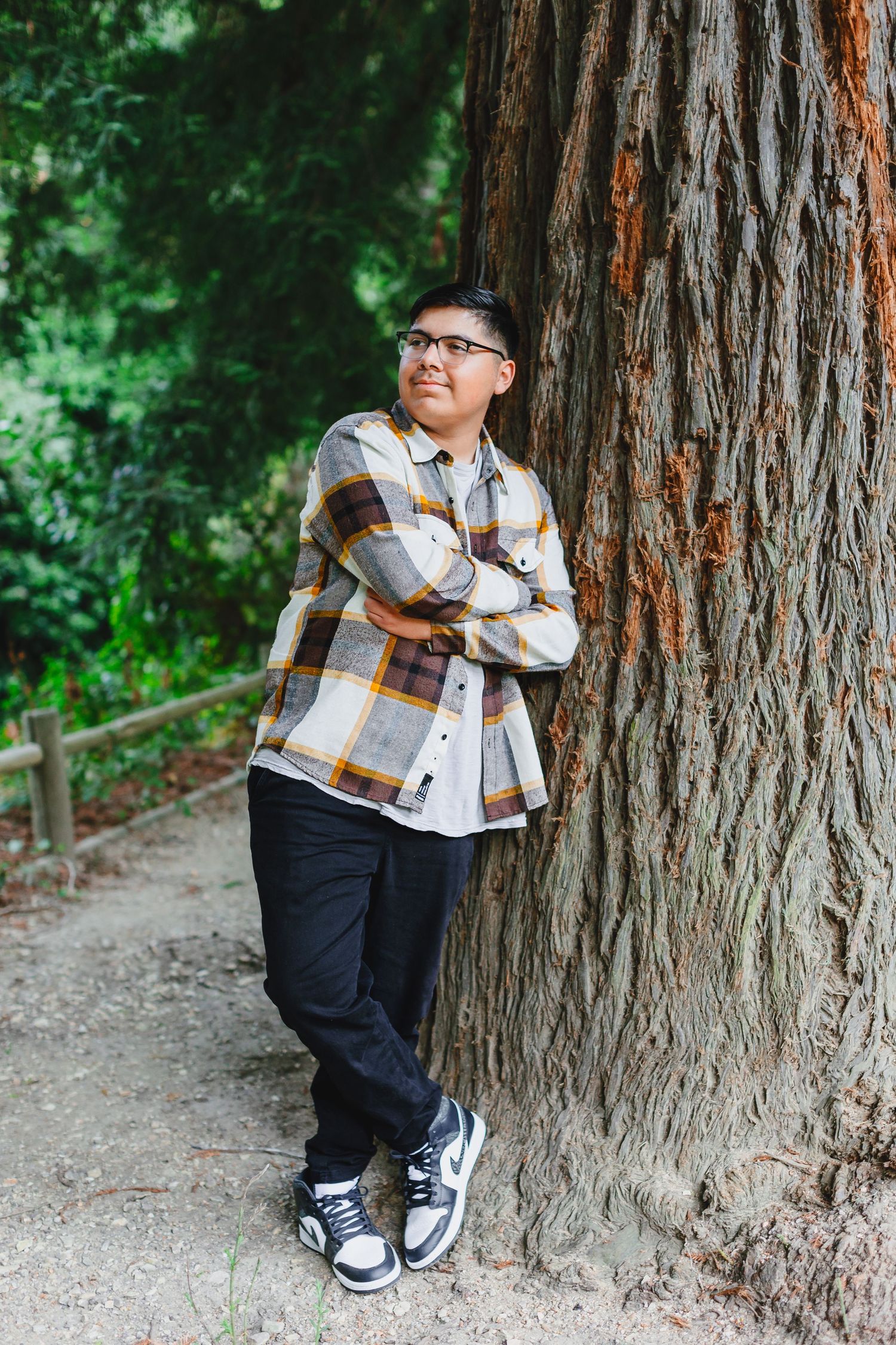 Young man in plaid flannel shirt and glasses leans against a large redwood tree in a forest path.