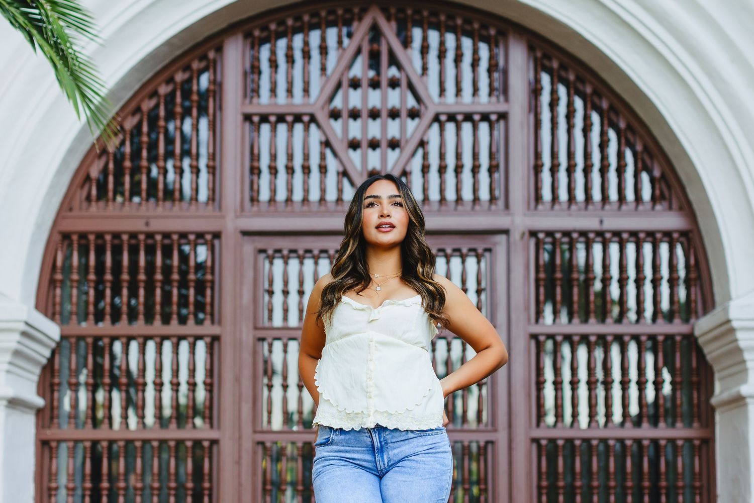 Young woman in white top and jeans poses confidently in front of ornate arched wooden door.