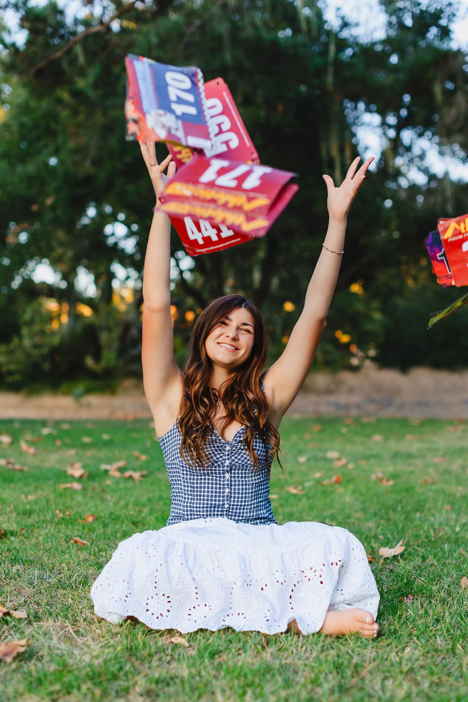 Young woman sitting on grass in white skirt, joyfully tossing colorful sale signs in the air outdoors.