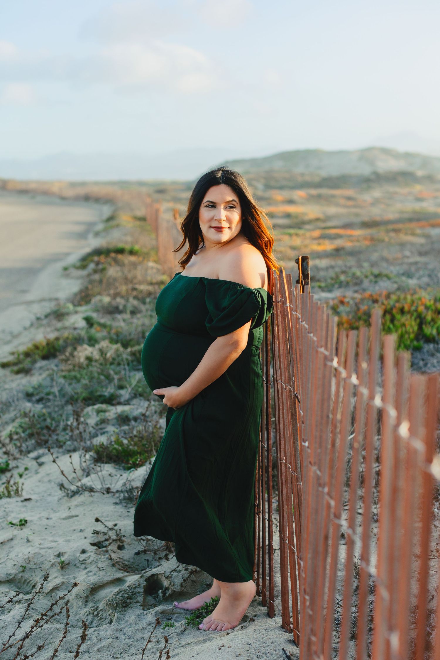 Beachside maternity portrait with rustic fence and coastal dunes.
