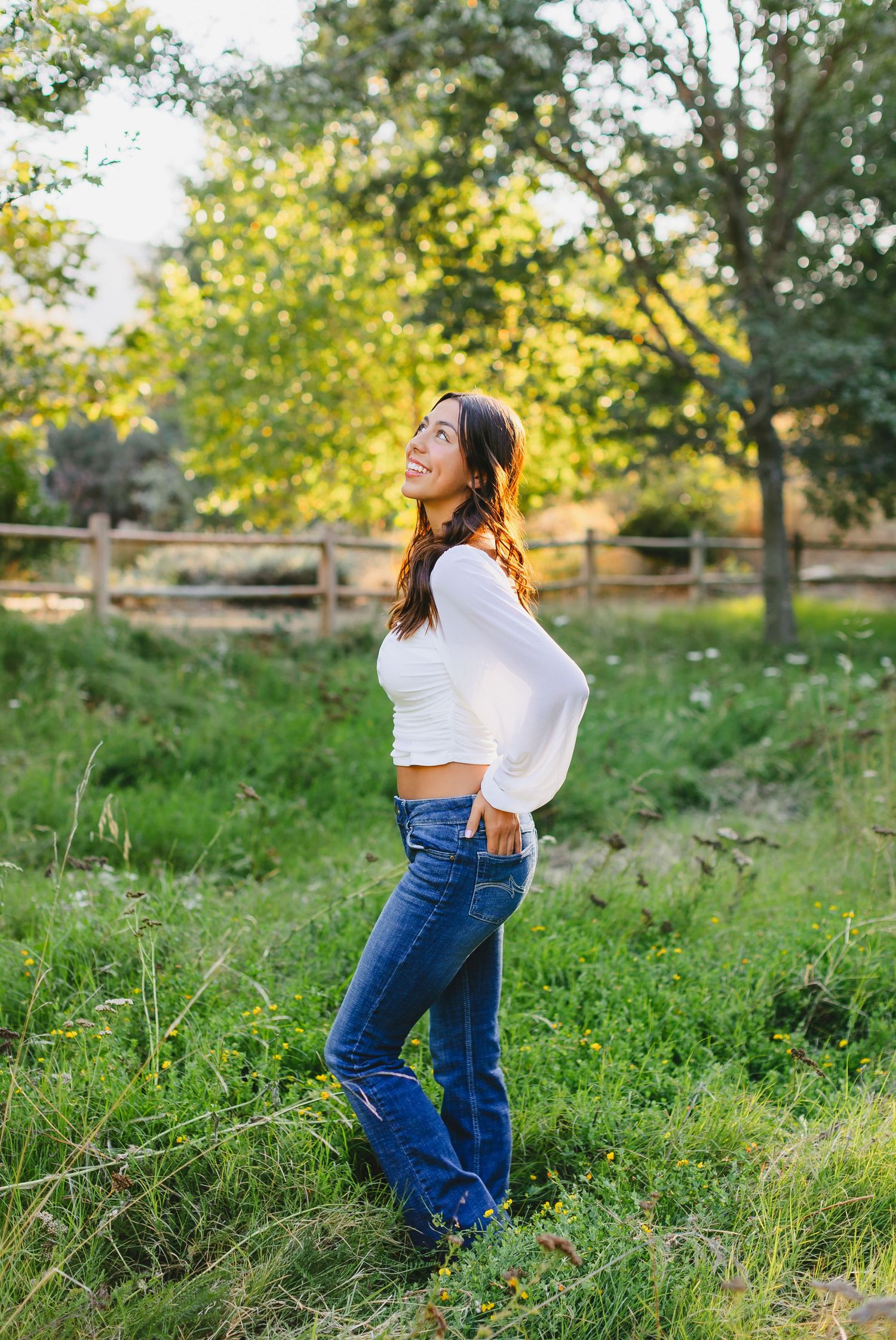 Person in white crop top and jeans posing in field with wooden fence and trees.
