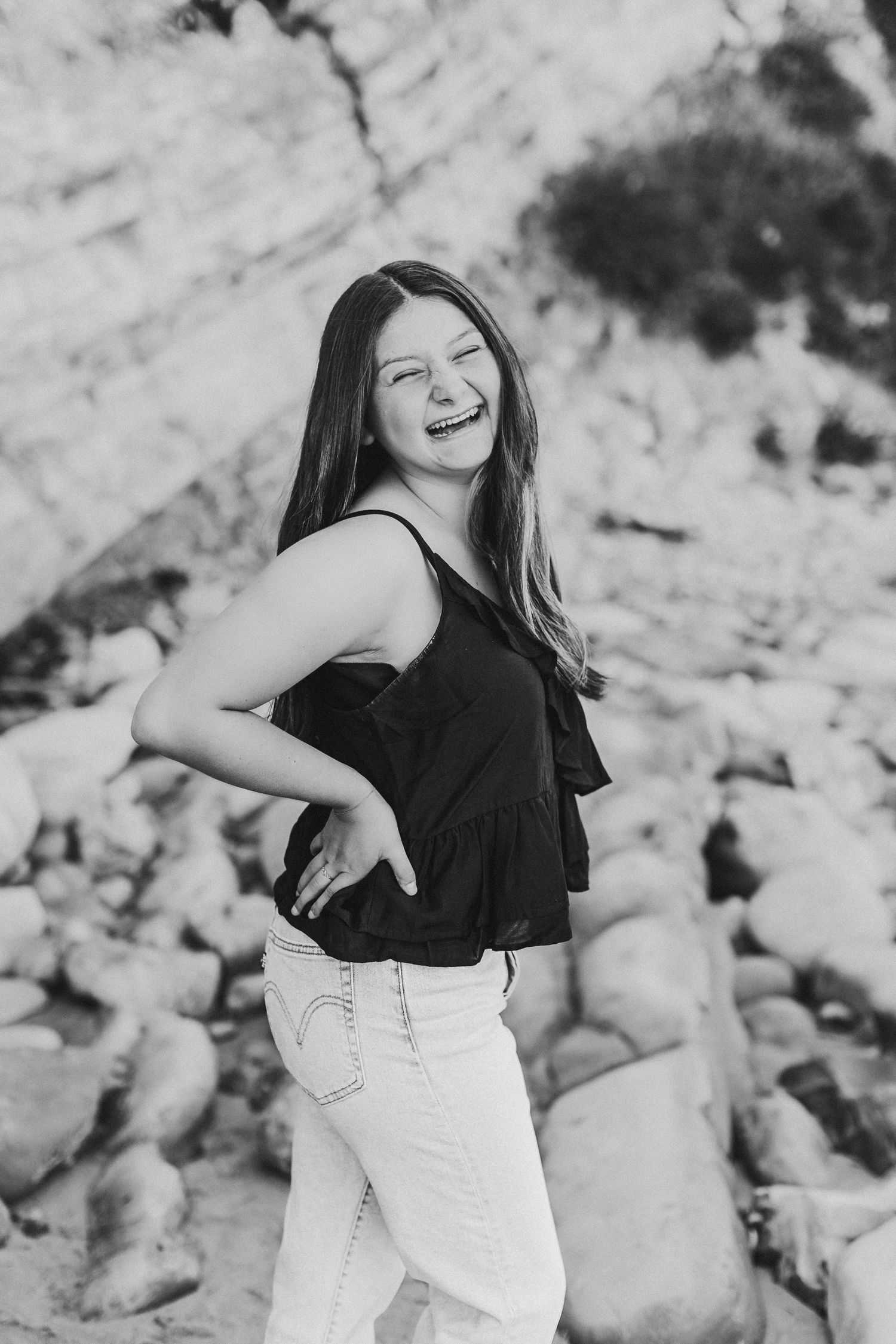 Black and white photo of a laughing young woman in a black top and white jeans posing against rocky background.