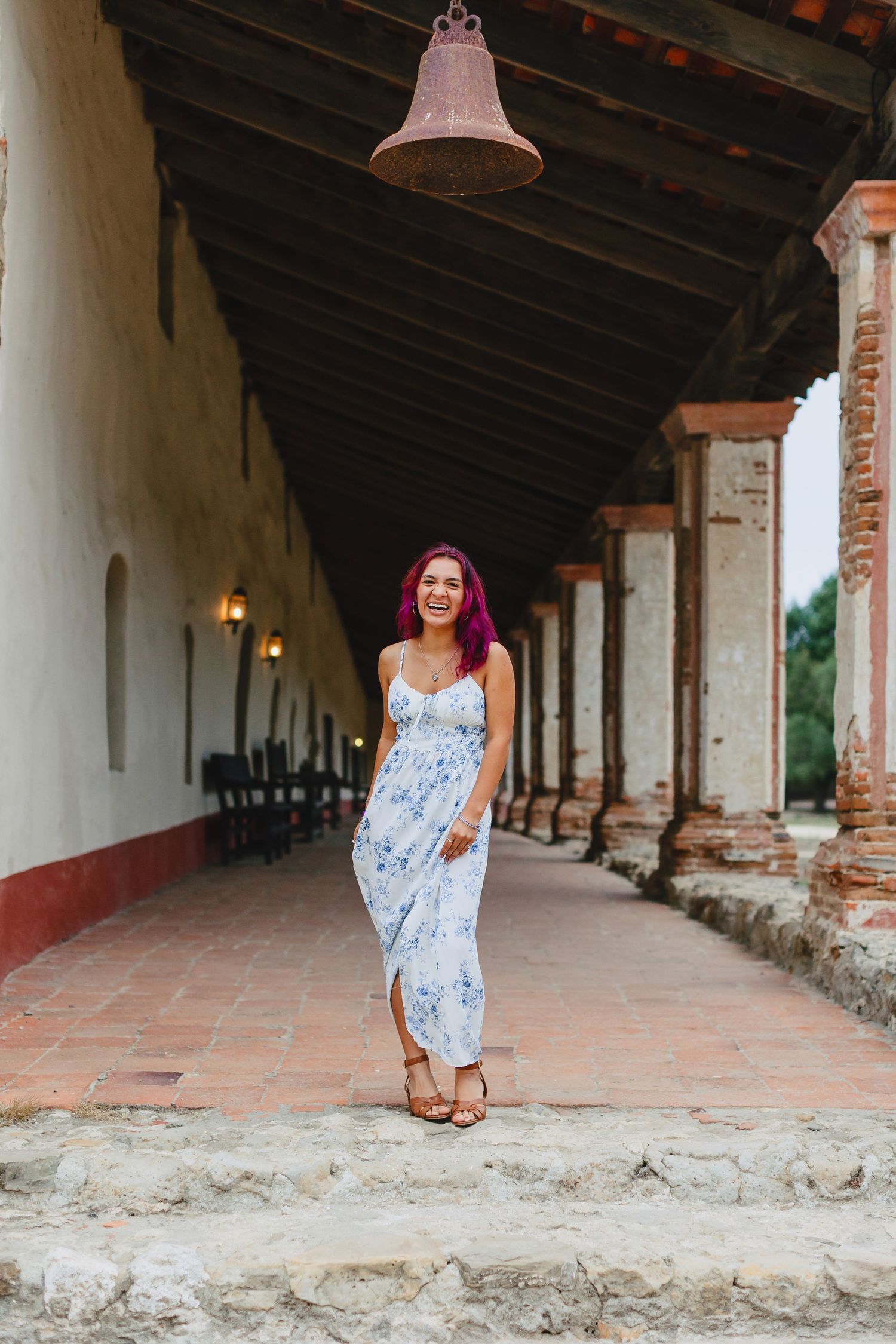 Woman with pink hair in a blue floral maxi dress stands smiling in a historic Spanish mission corridor.