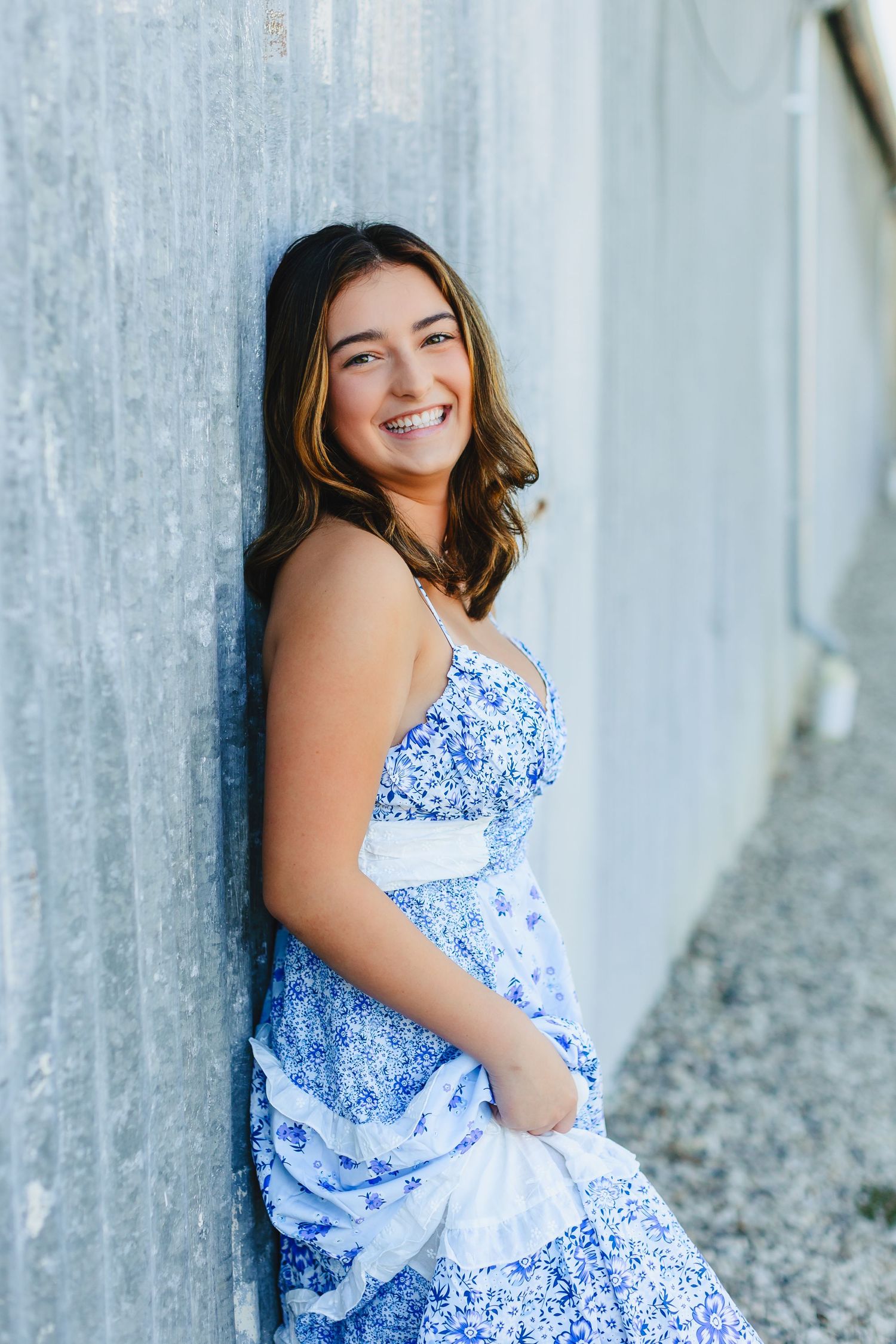 Smiling teenage girl in blue floral sundress leaning against concrete wall outdoors.