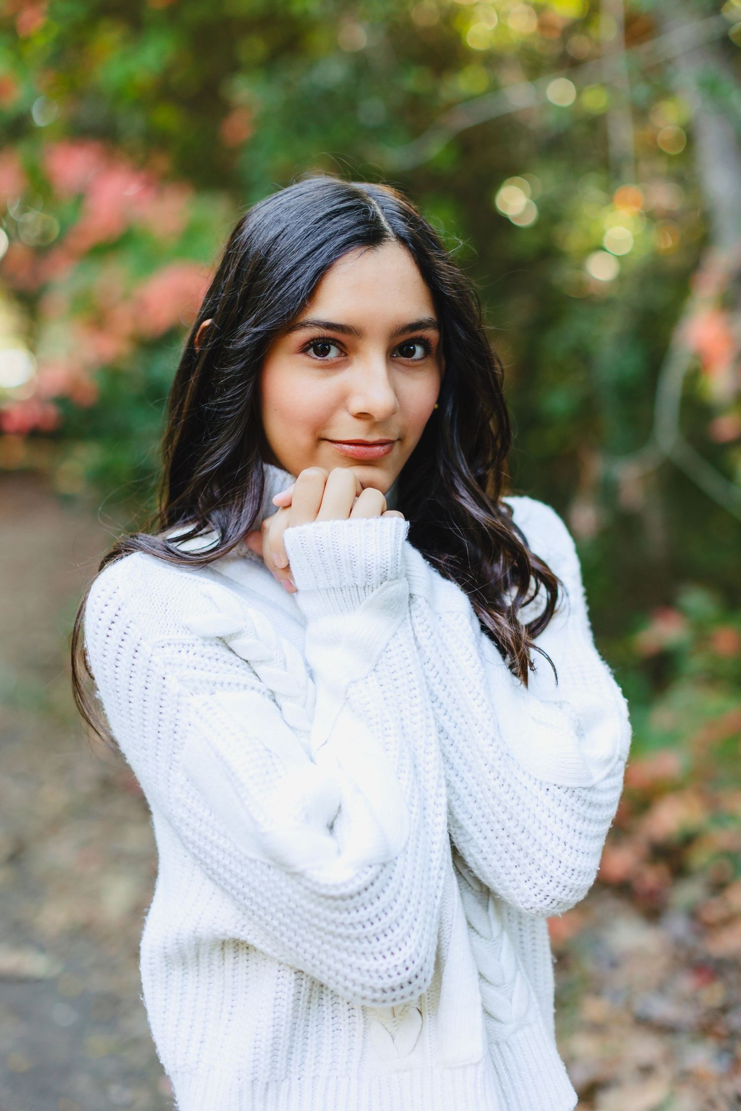 Young woman in white cable-knit sweater posing outdoors among colorful autumn foliage.