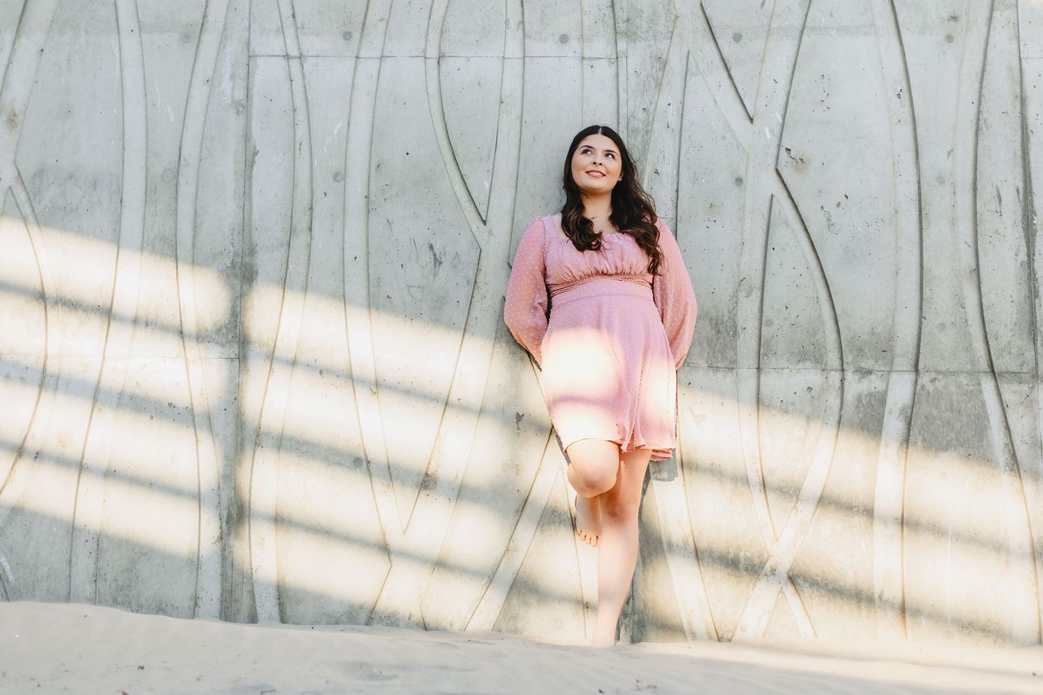 A woman in a pink dress leans against a concrete wall, bathed in soft sunlight and shadow patterns.