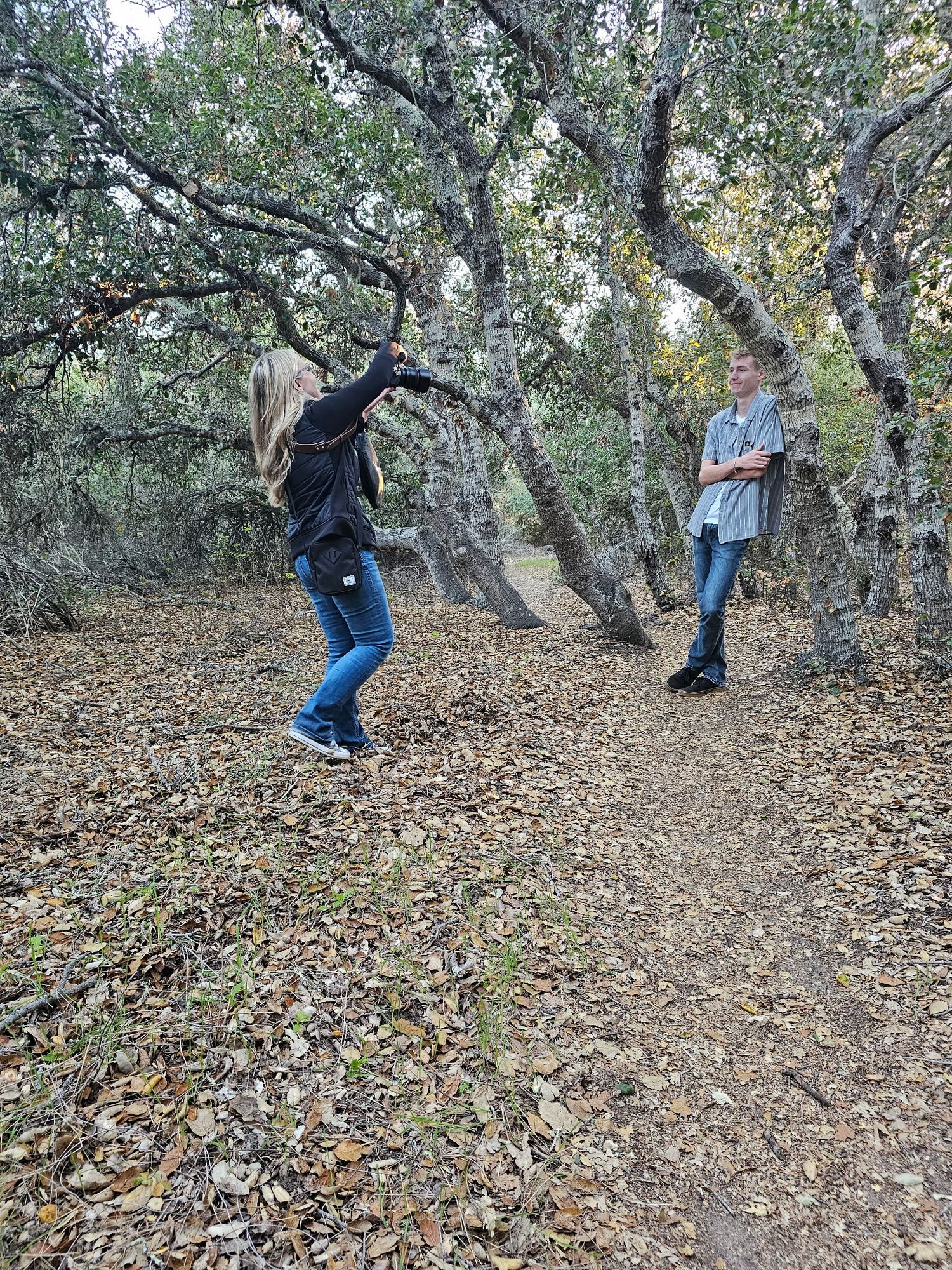 Two people among twisted trees on leaf-covered forest trail in autumn.