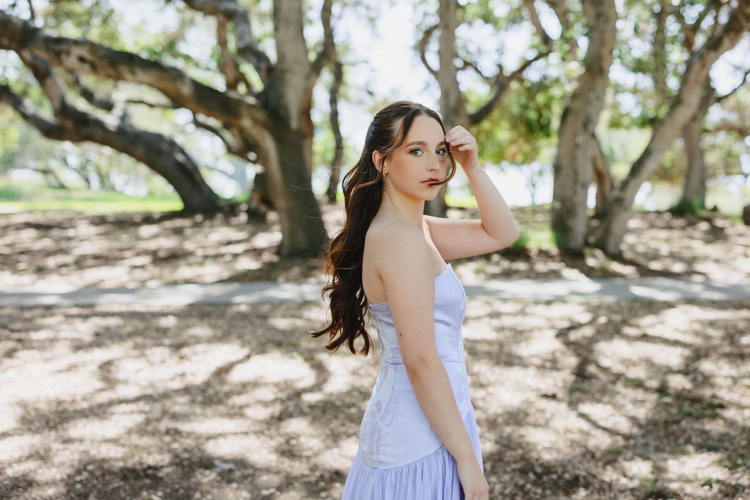 Young woman in white sundress poses gracefully among large trees in a sunlit park setting.