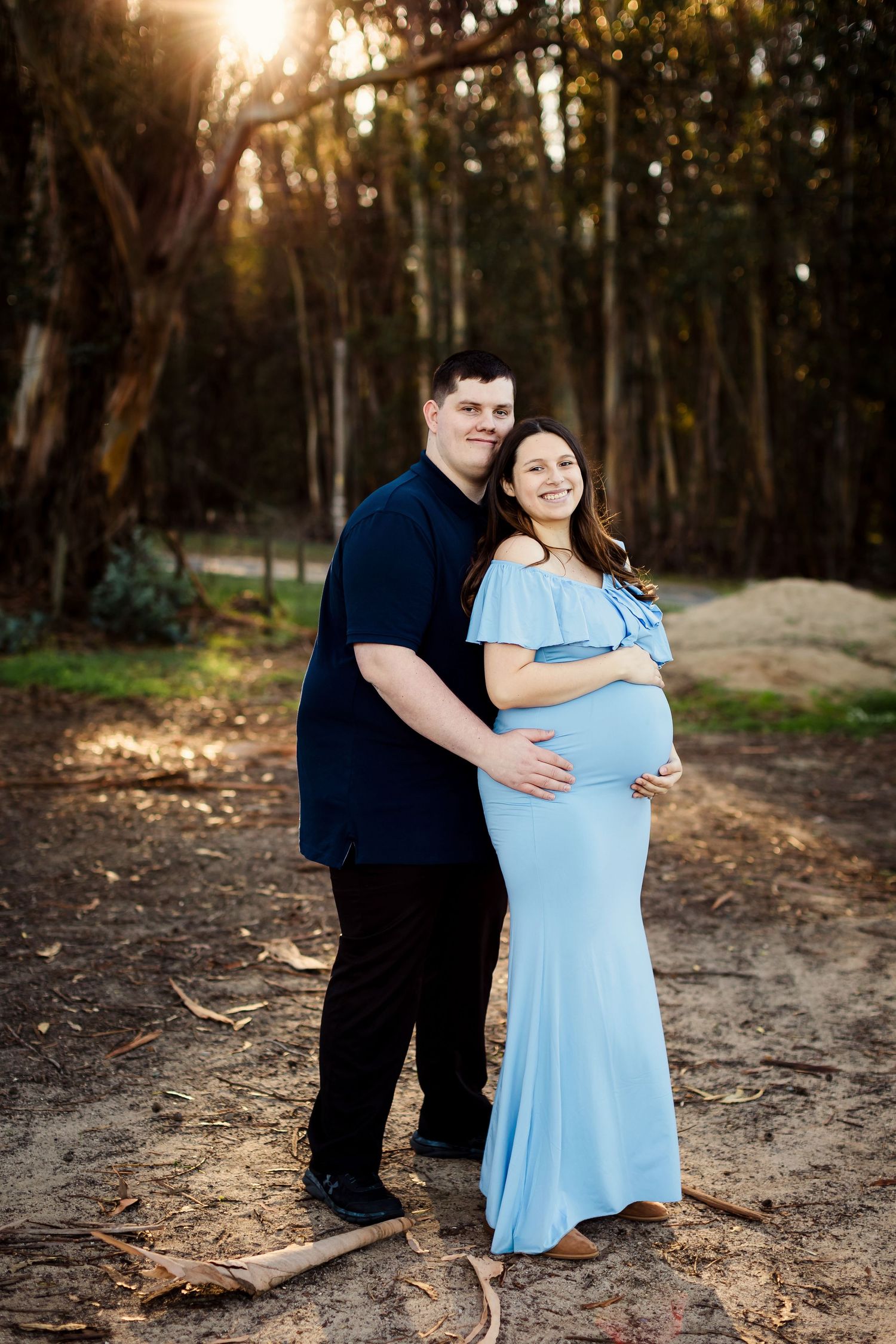 A couple embraces in a sunlit forest setting while expecting a baby, wearing a light blue maternity dress and dark clothing.