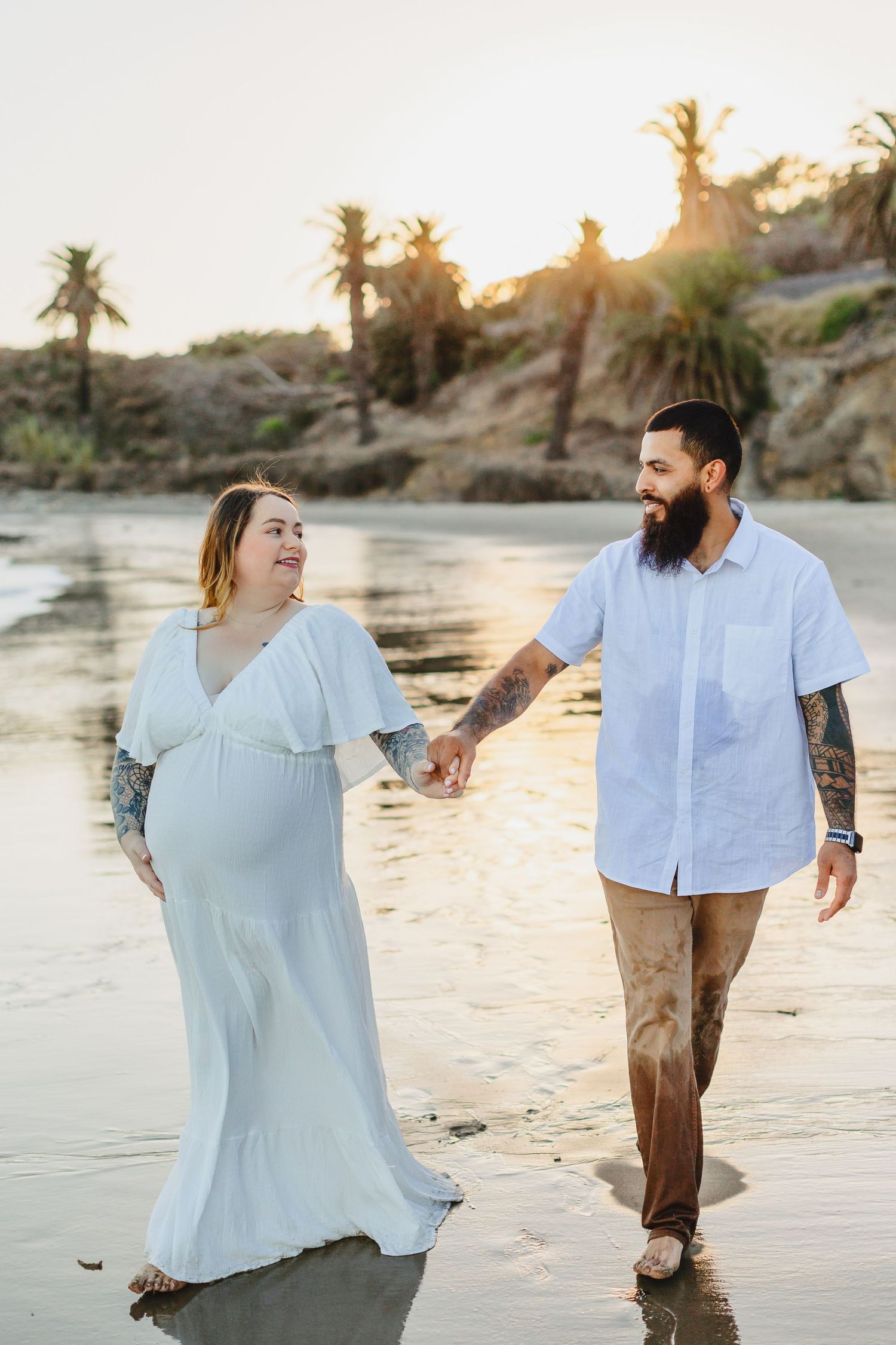 Couple walking hand in hand on beach at sunset with palm trees in background.