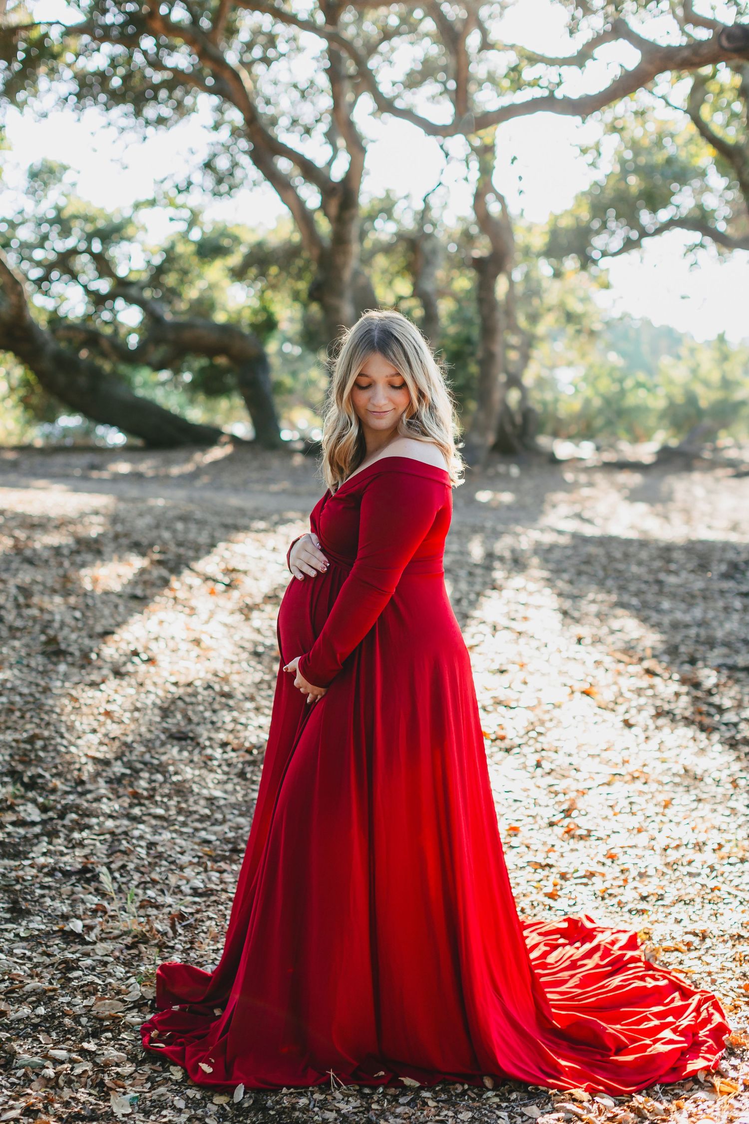 A flowing red velvet maternity gown catches sunlight beneath oak trees in an outdoor photoshoot.