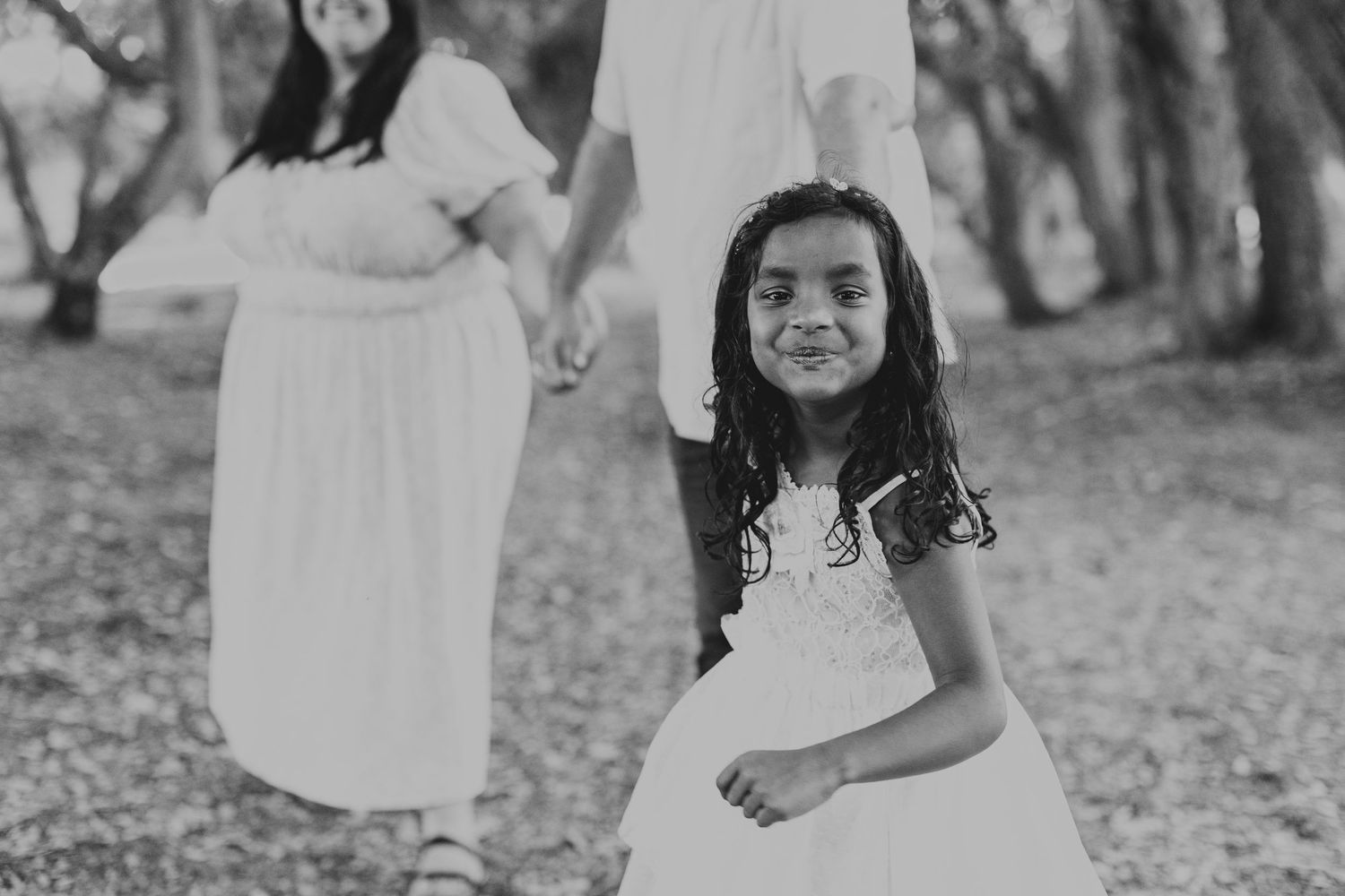 Black and white photo of a young child wearing a white dress standing in an outdoor wooded setting.