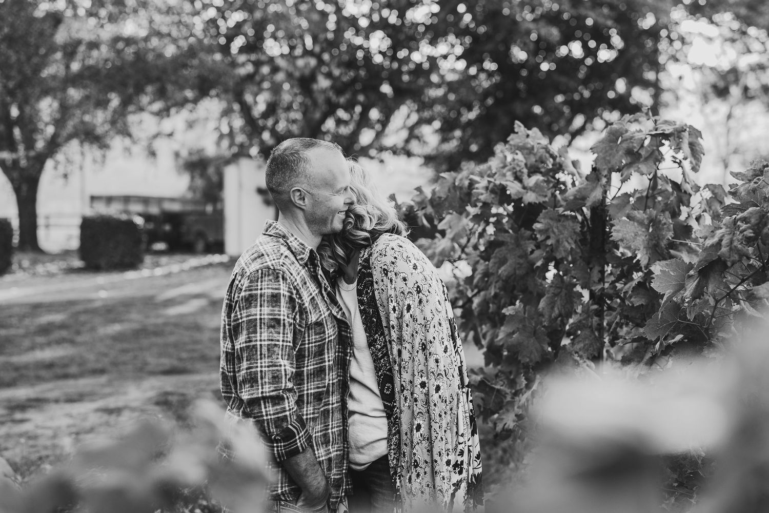 A couple in plaid shirts embraces during an outdoor photo session in a rustic wooded setting.