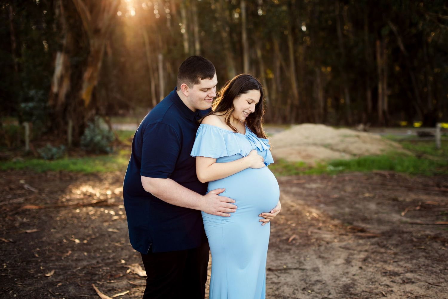 Maternity photoshoot in a sunlit forest with a couple in casual attire embracing her baby bump.