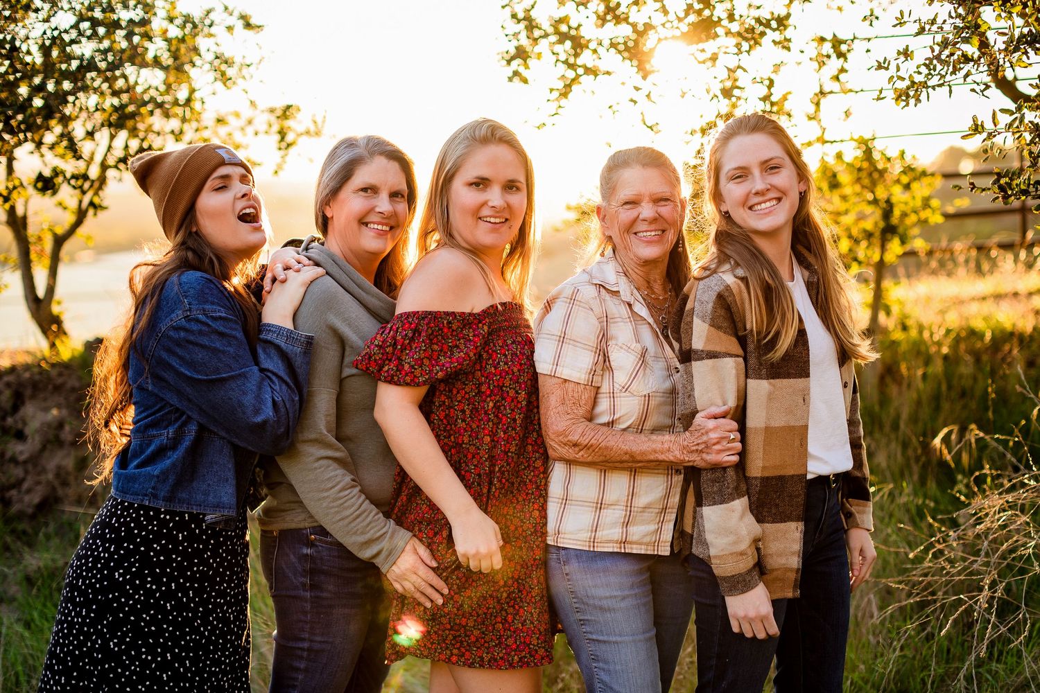Group of people laughing and embracing during golden hour sunset in a field with tall grass and warm lighting.