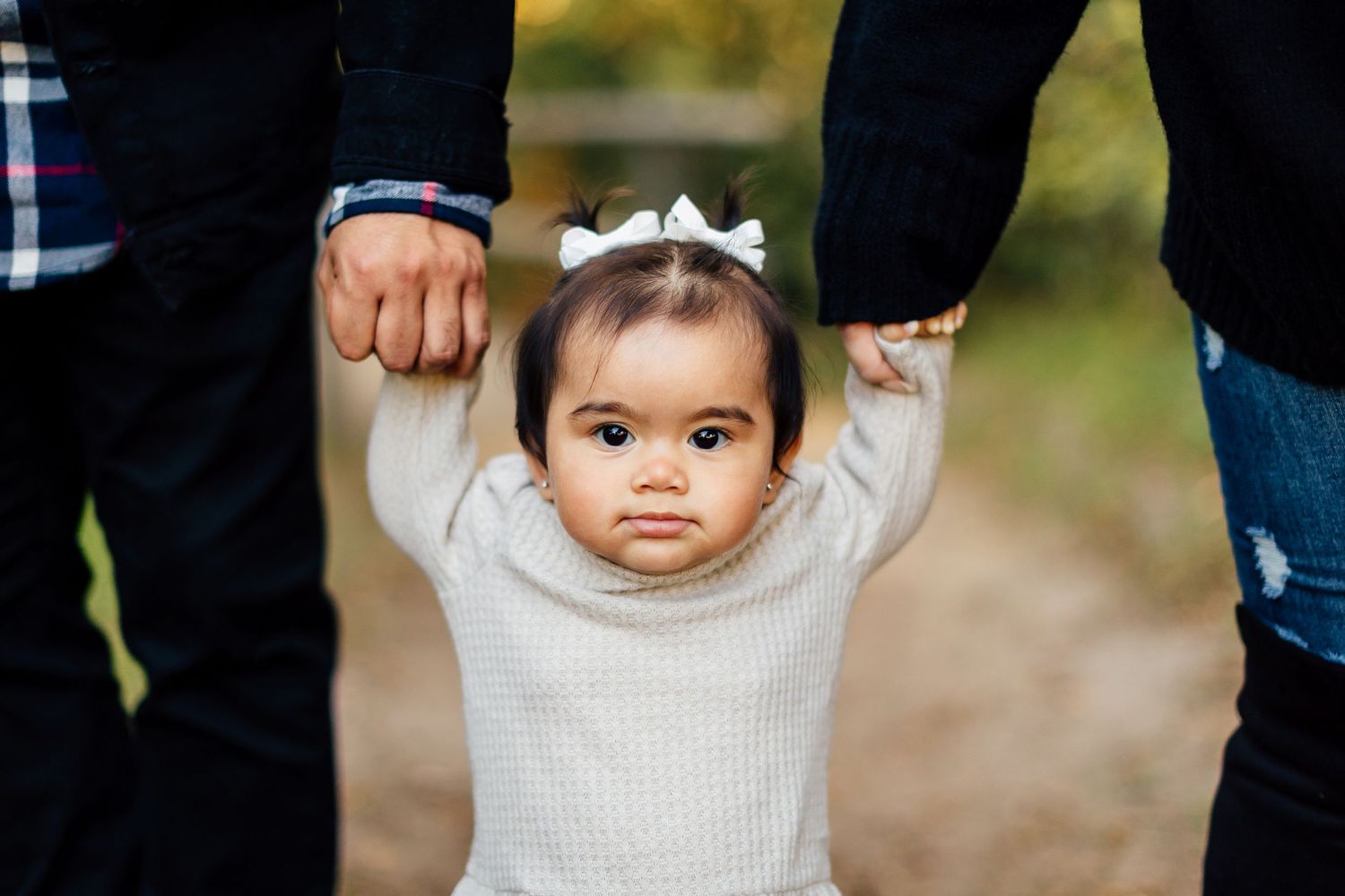 A child stands between parents' legs holding hands in an outdoor setting during autumn.