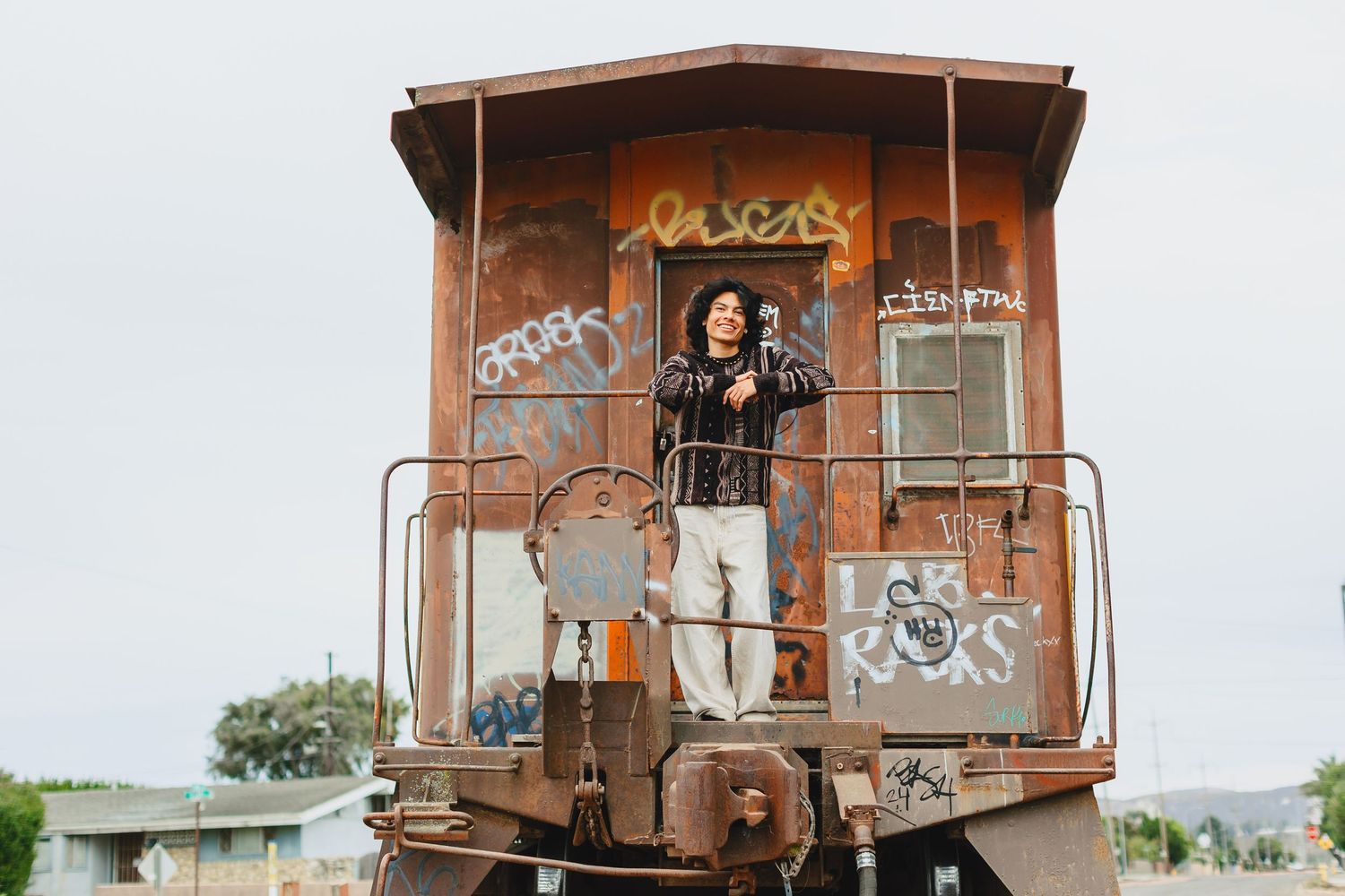 Young person standing on vintage graffiti-covered train caboose holding camera outdoors.