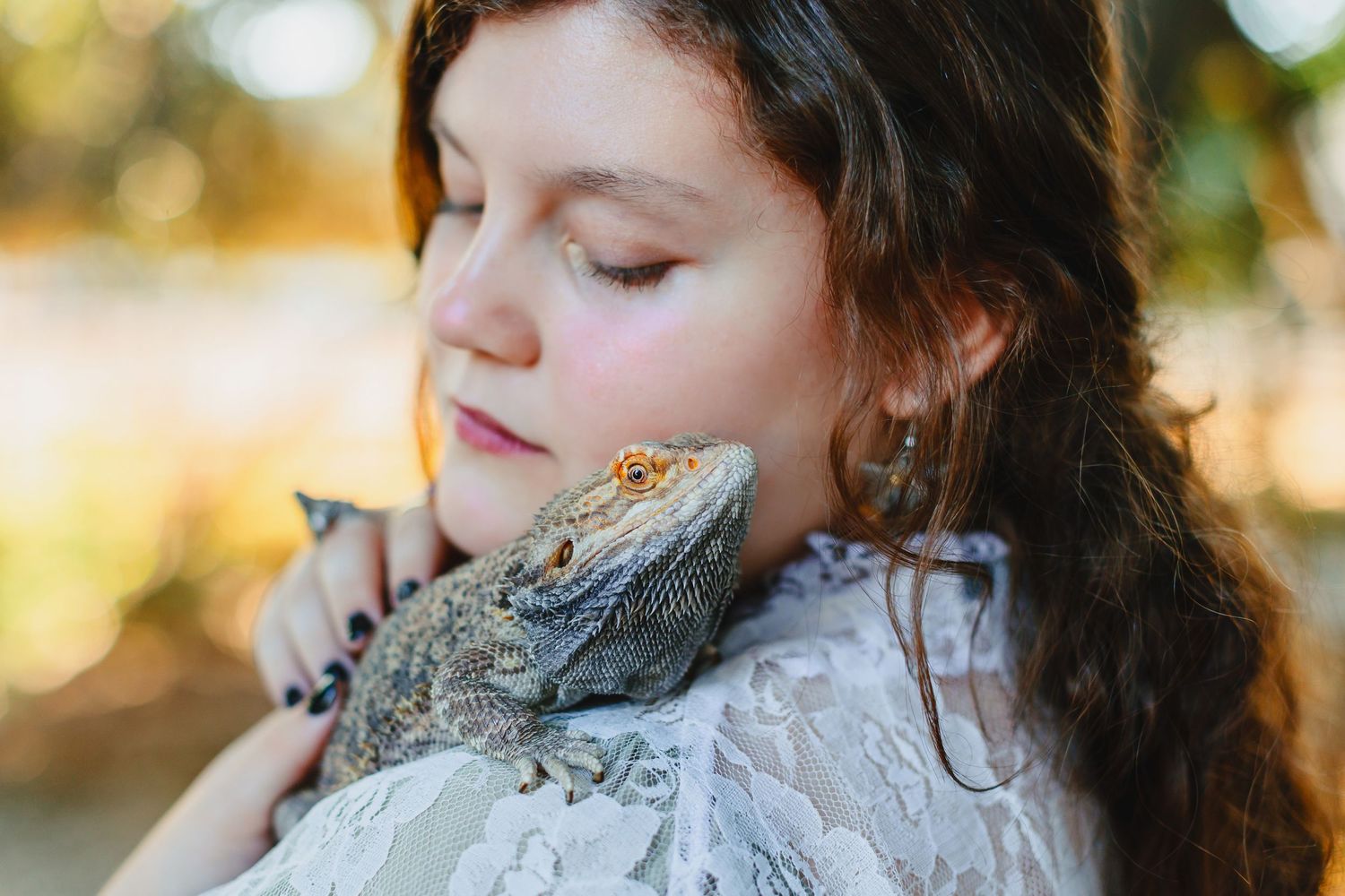 Young girl with closed eyes tenderly holding a bearded dragon lizard on her shoulder outdoors.