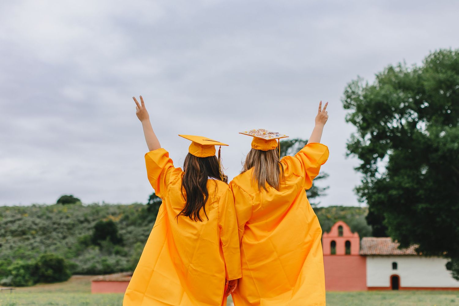 Two graduates in bright yellow caps and gowns raise peace signs while facing away from the camera on graduation day.
