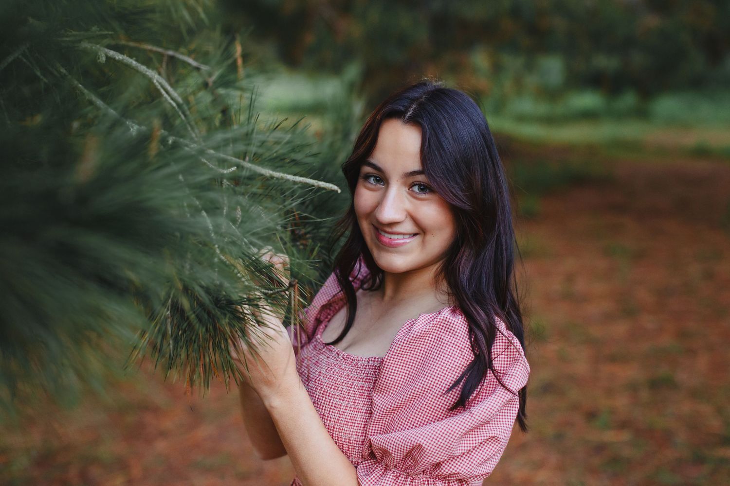 Someone in a pink dress poses near pine branches outdoors with a warm smile on a sunny day.