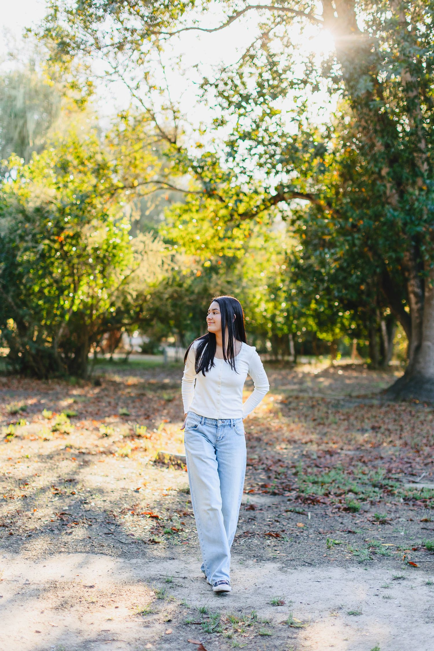 Young woman in white sweater and light blue jeans walking on a sunny park path surrounded by green trees.