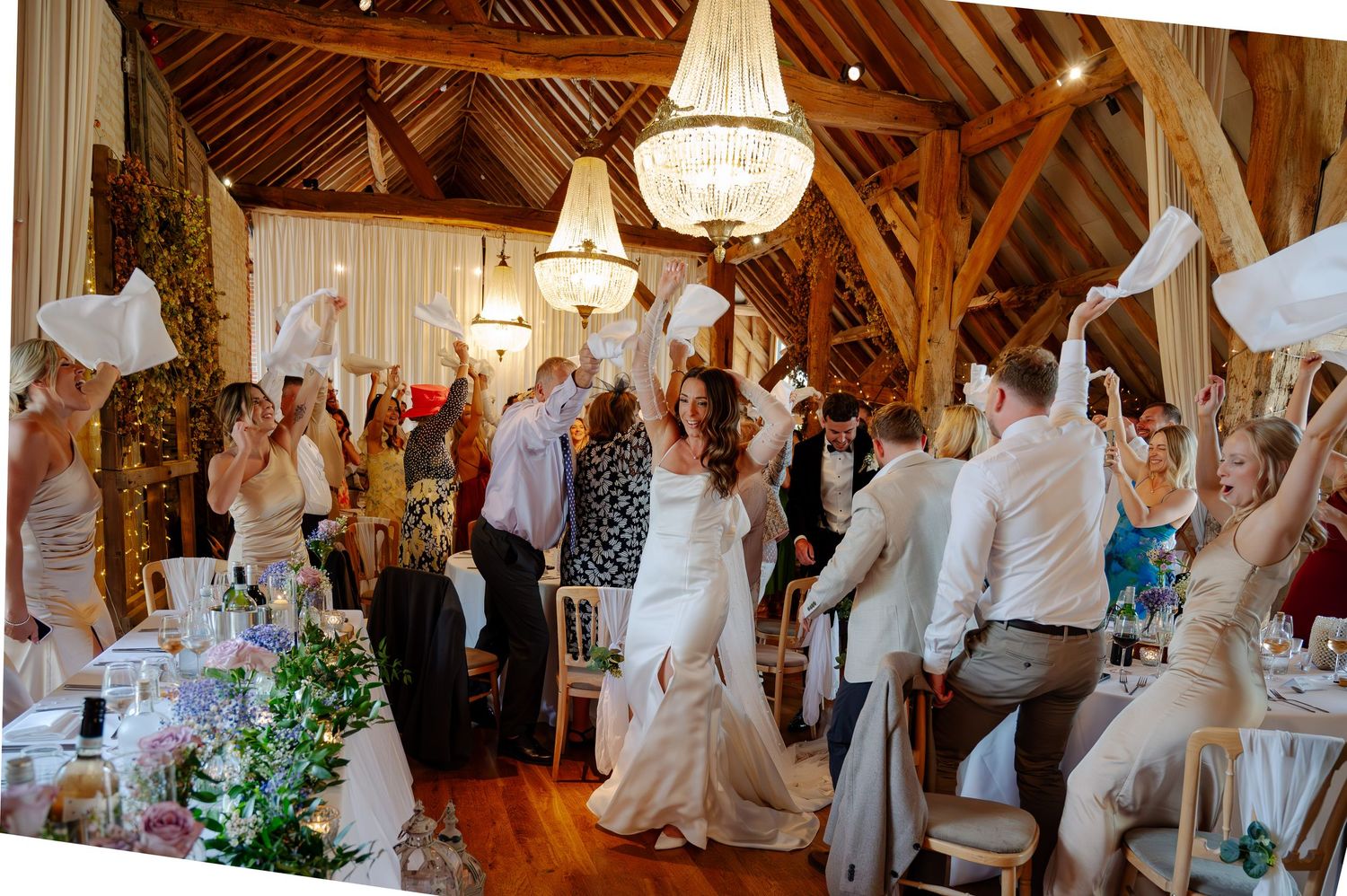 Wedding guests celebrate during a reception in a rustic barn venue with elegant chandeliers and exposed wooden beams.