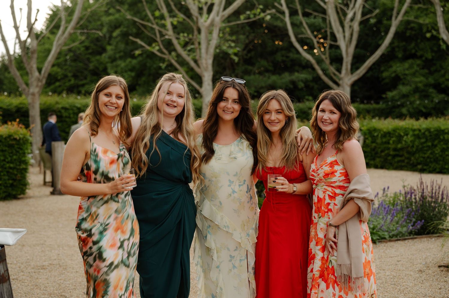 A group of people in formal attire pose together outdoors for wedding photos in a garden setting.