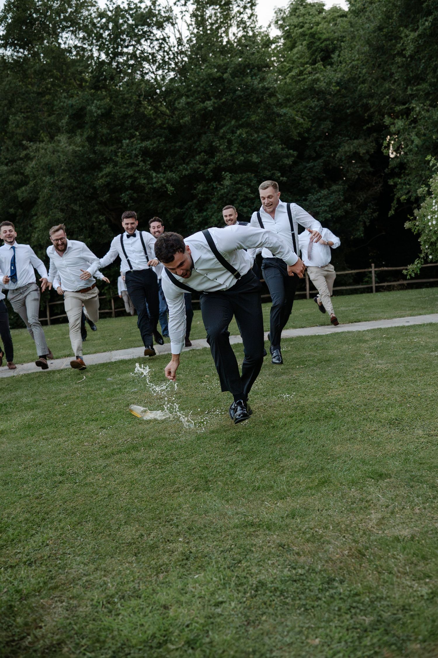 Groomsmen in white shirts and suspenders perform a fun jumping photo sequence on a grassy lawn.