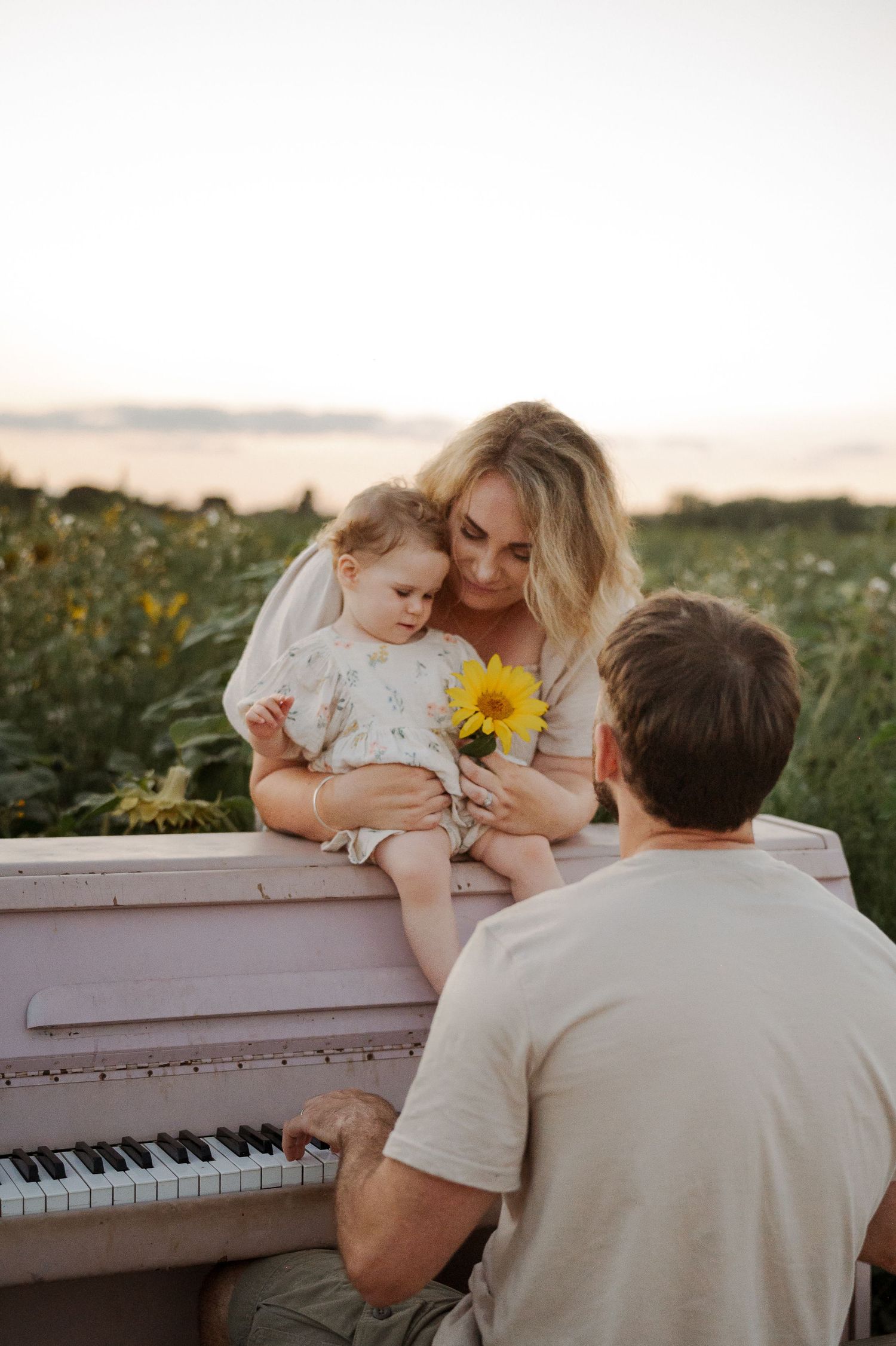 Family with baby girl holding sunflower on pink piano in field at sunset.