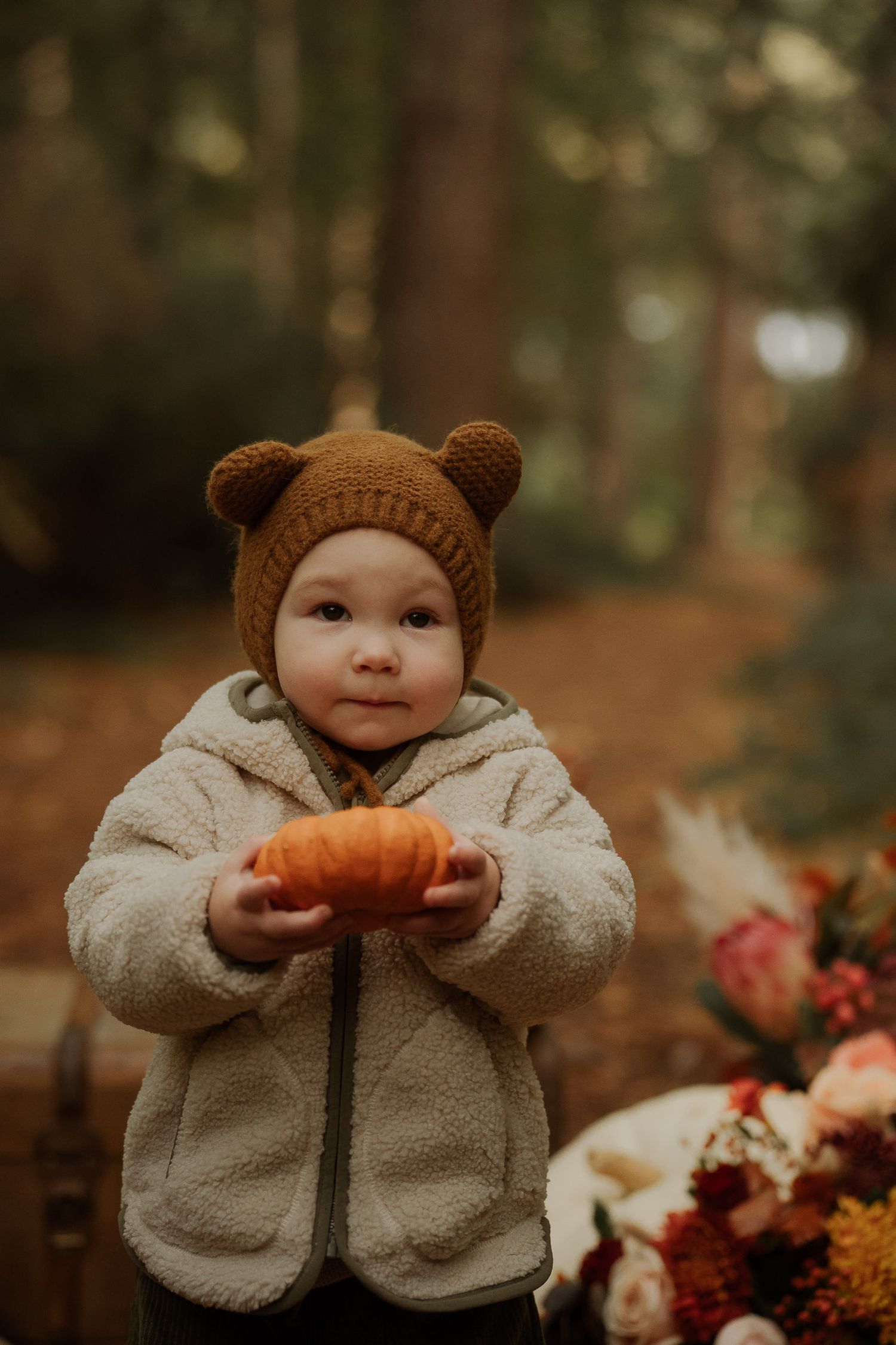 Adorable baby in bear hat holding small pumpkin in autumn forest setting with fall flowers.
