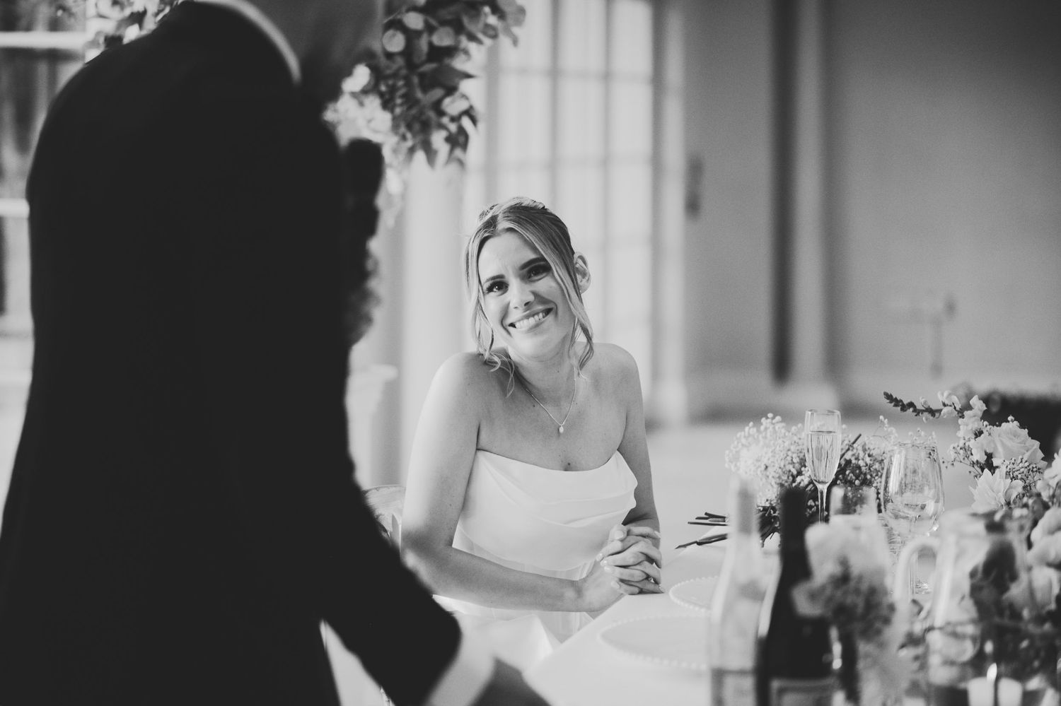 Black and white photo of a smiling bride in a strapless gown seated at a wedding reception table.