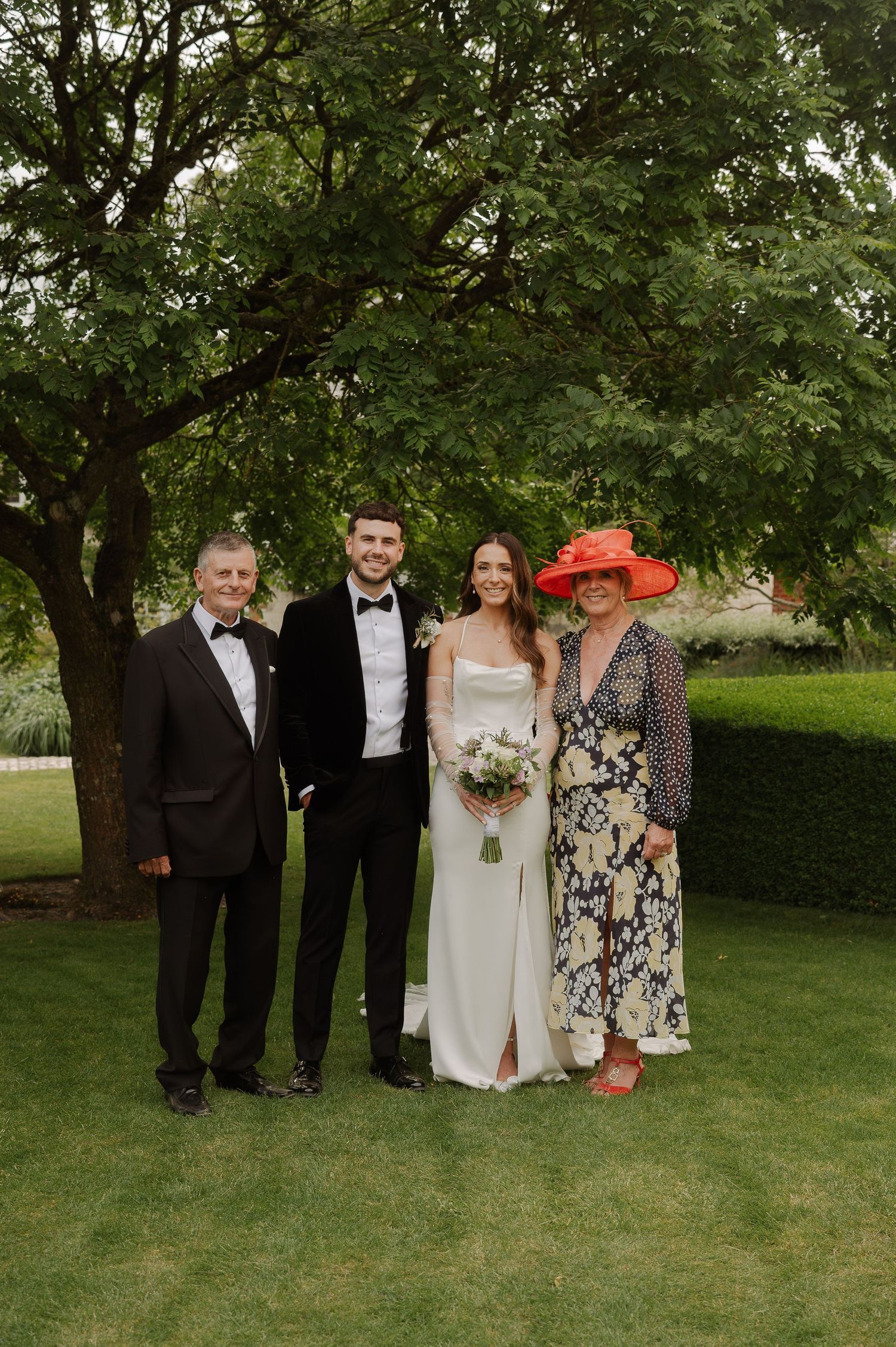 Wedding party group photo taken outdoors on a green lawn with trees in the background.