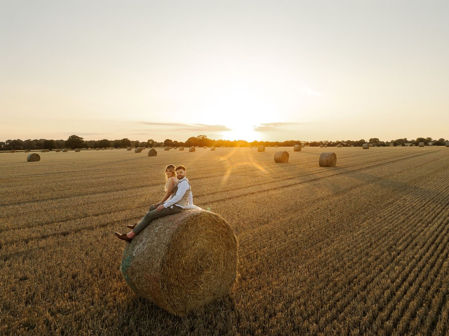 A couple embraces in a golden wheat field at sunset while hay bales dot the rural landscape.