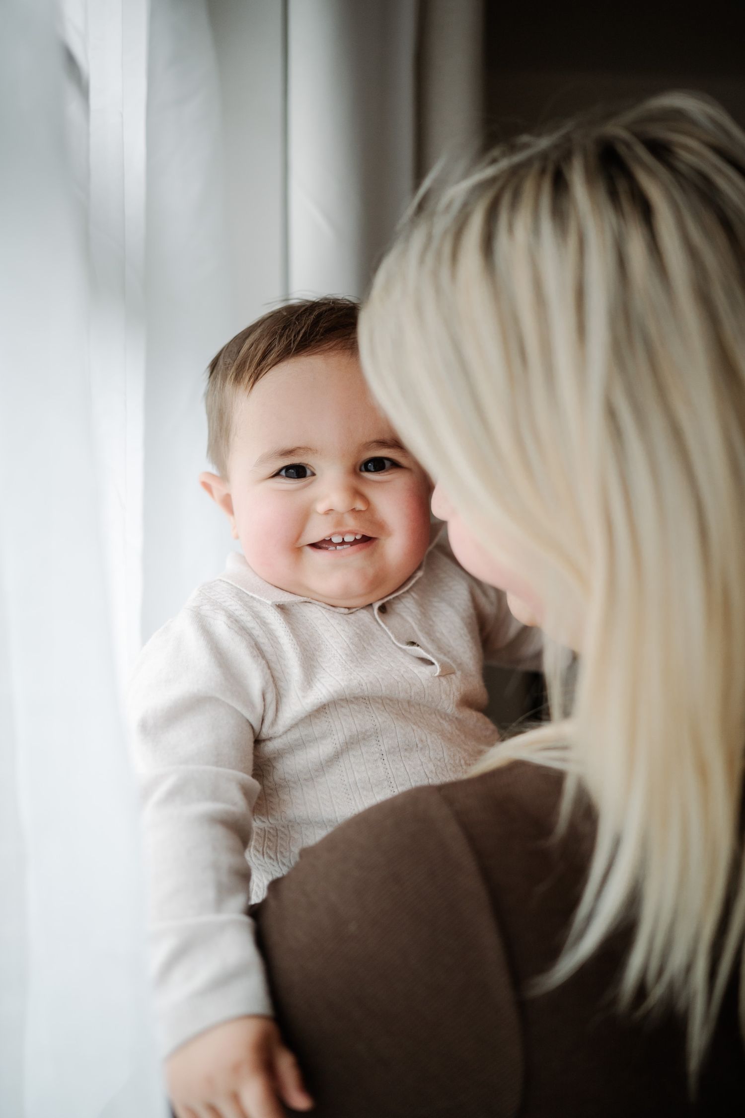 A smiling baby in a white onesie being held by a blonde woman near a window in a tender moment.