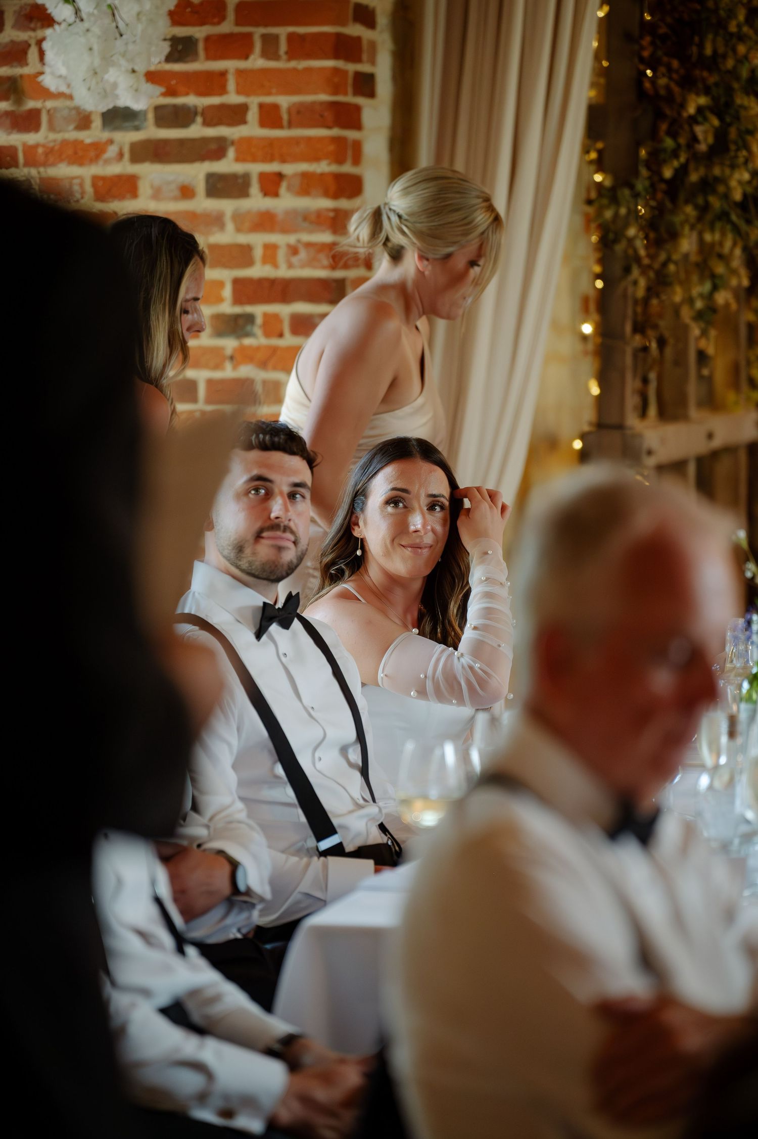 Wedding reception guests seated at tables with exposed brick wall and string lights in background.