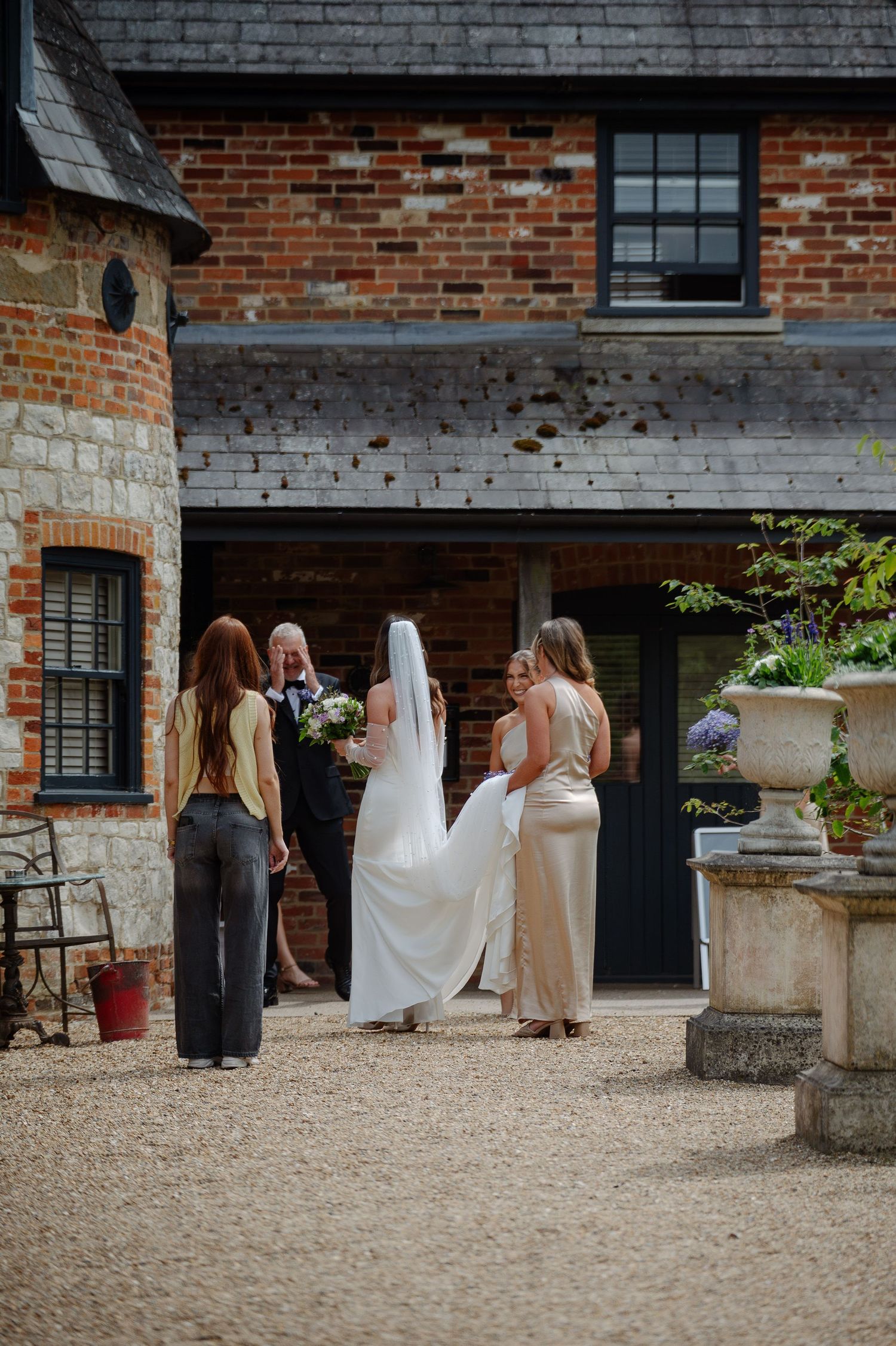 Wedding party gathers outside historic brick building with gravel courtyard.