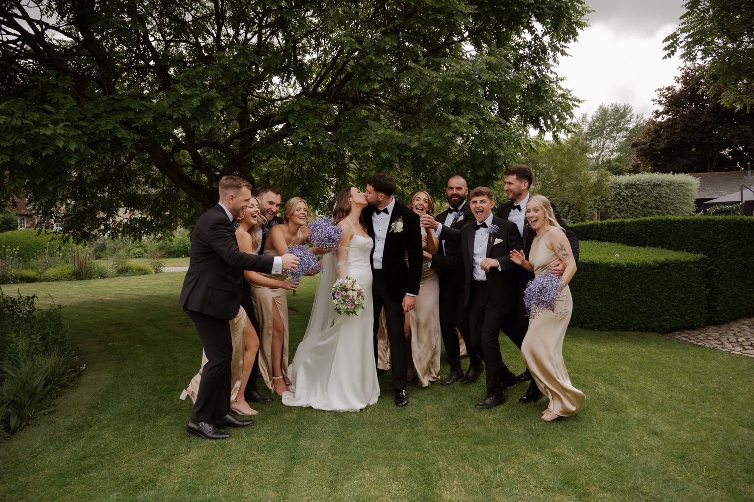 Wedding party poses playfully together on a green lawn during outdoor summer celebration.