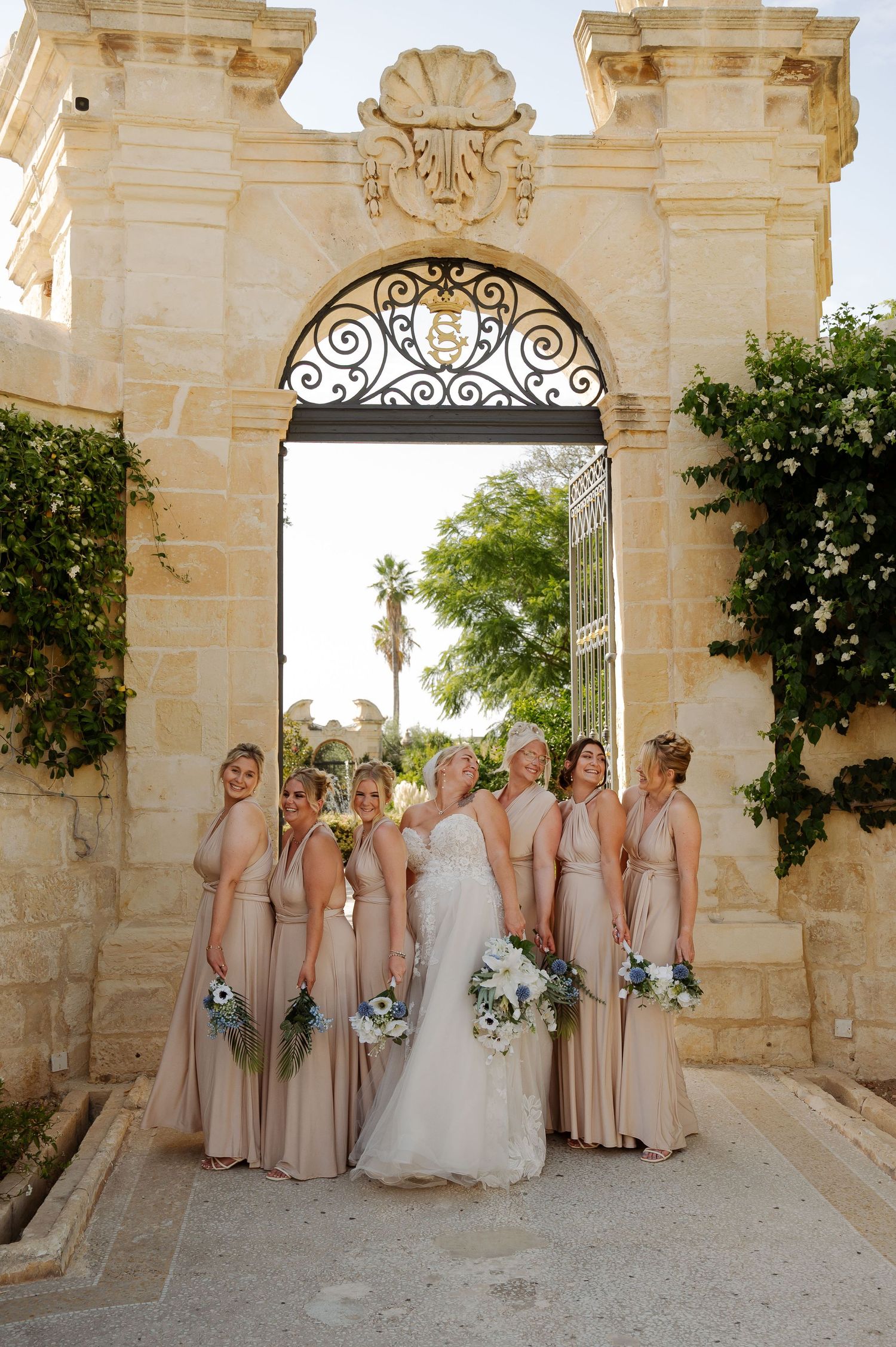 Wedding party poses under ornate stone archway with wrought iron details and palm trees visible in background.