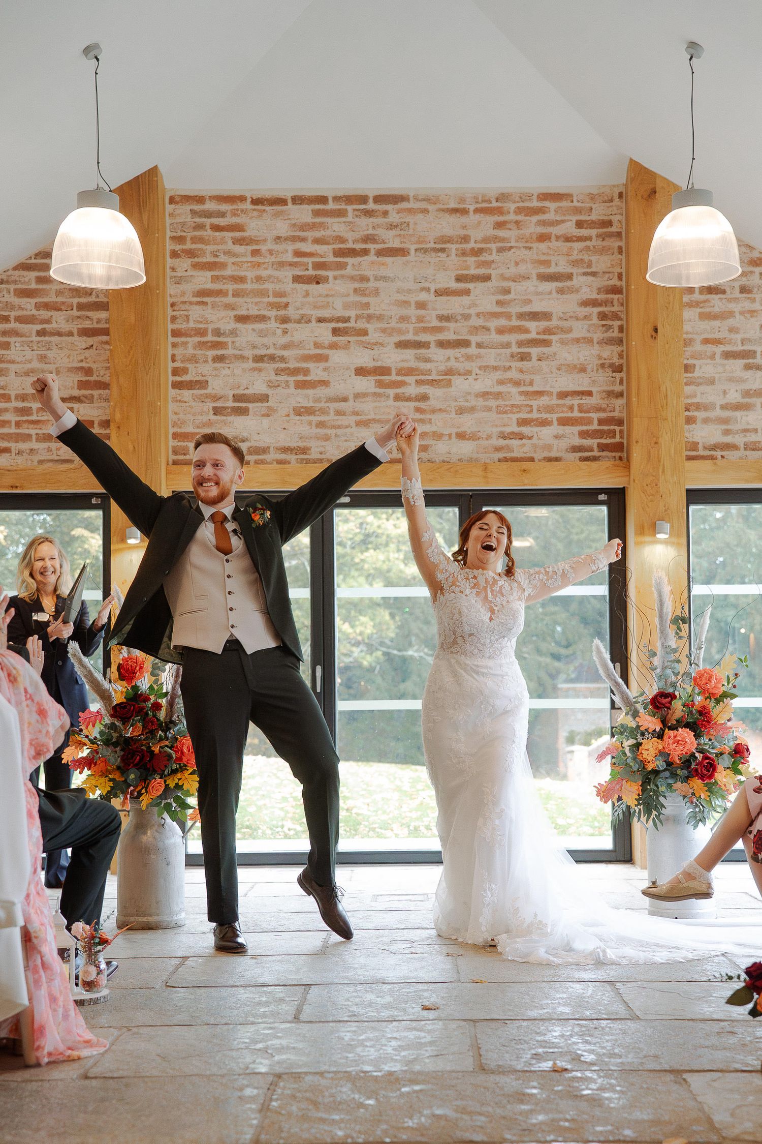 Joyful couple celebrating at their wedding reception with brick wall backdrop and floral arrangements.