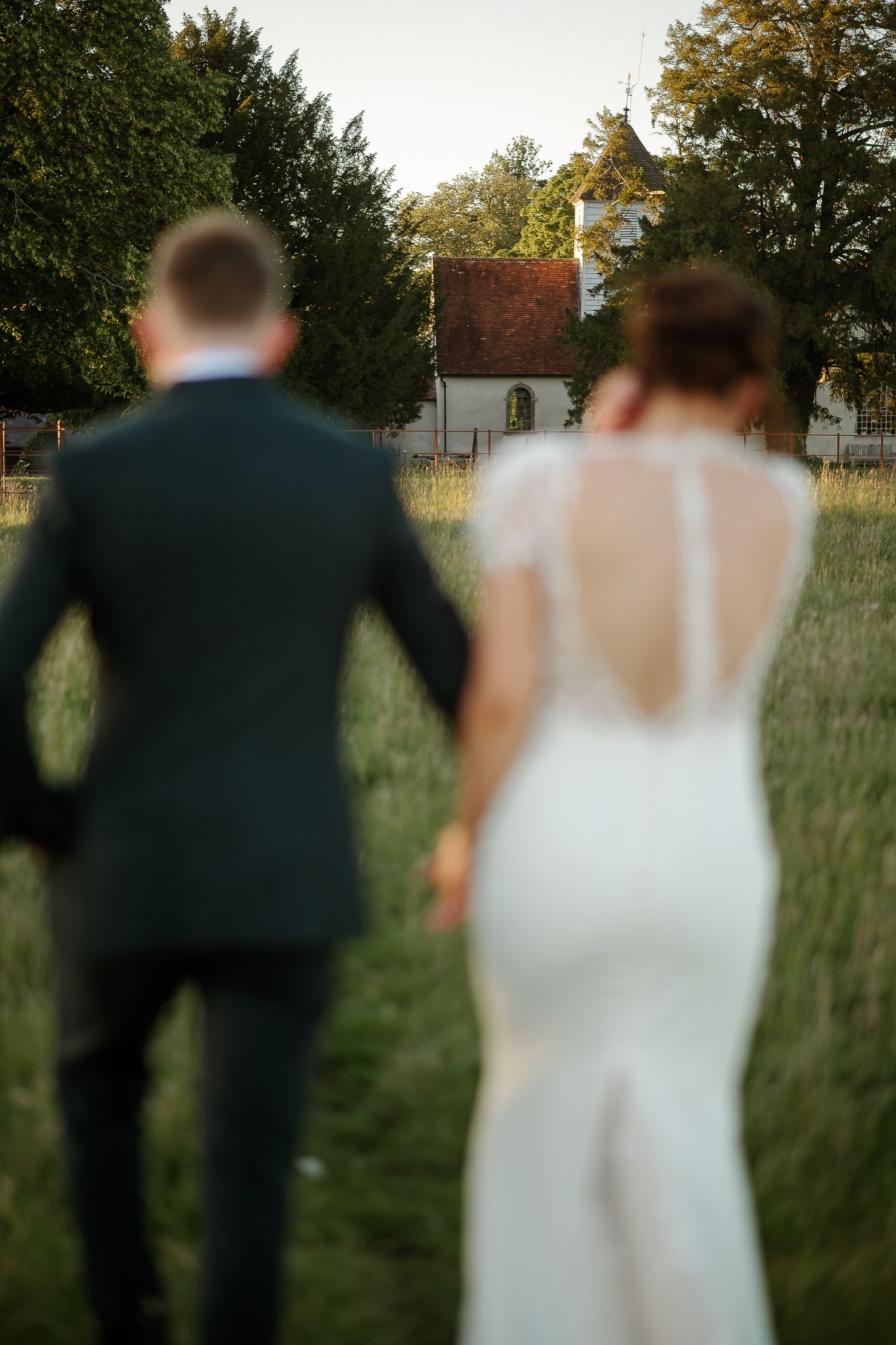 Bride and groom walking hand in hand through a green meadow toward a charming white chapel surrounded by trees.