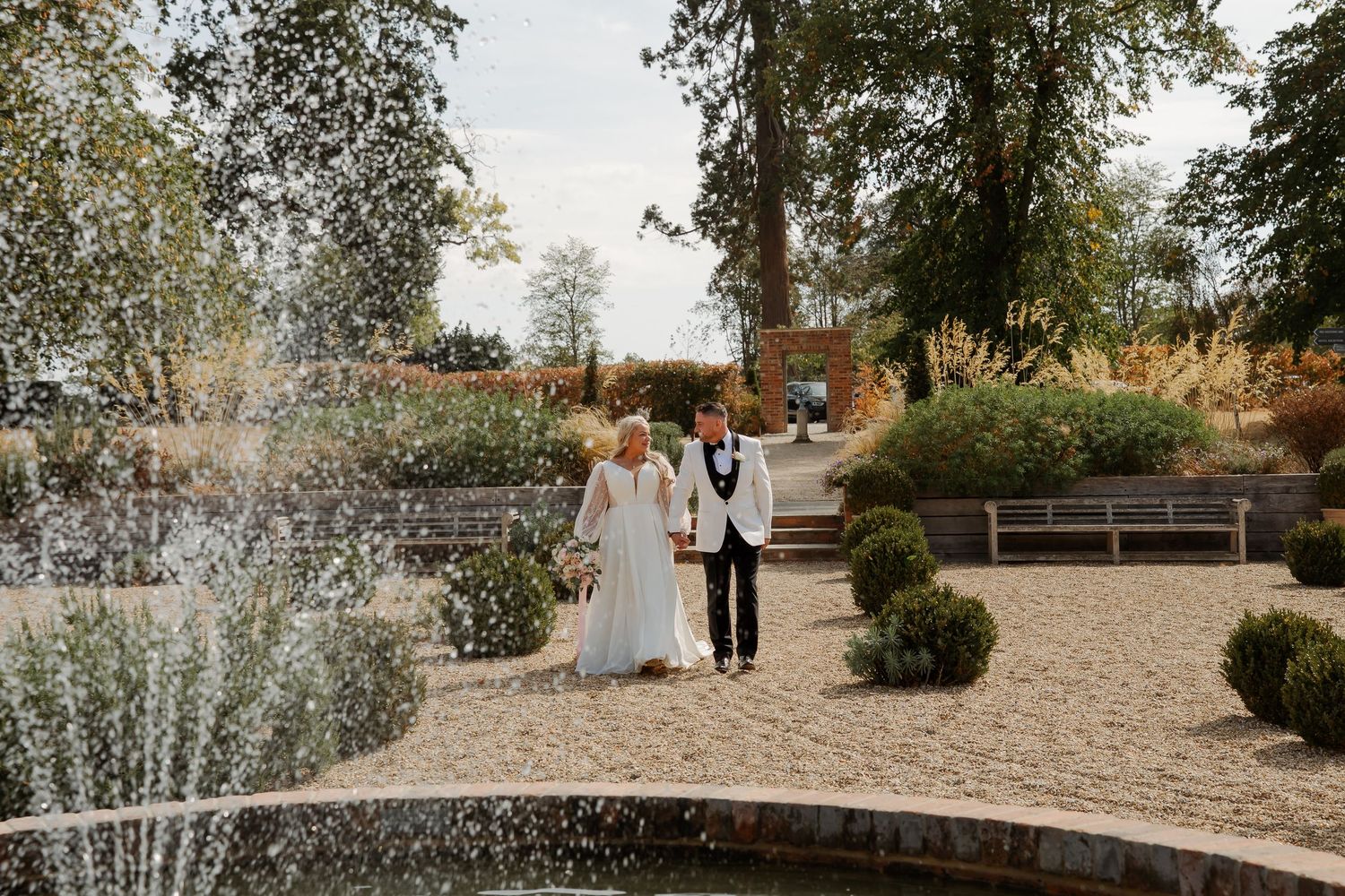 A bride and groom walk hand in hand near a fountain in a beautifully landscaped garden on their wedding day.