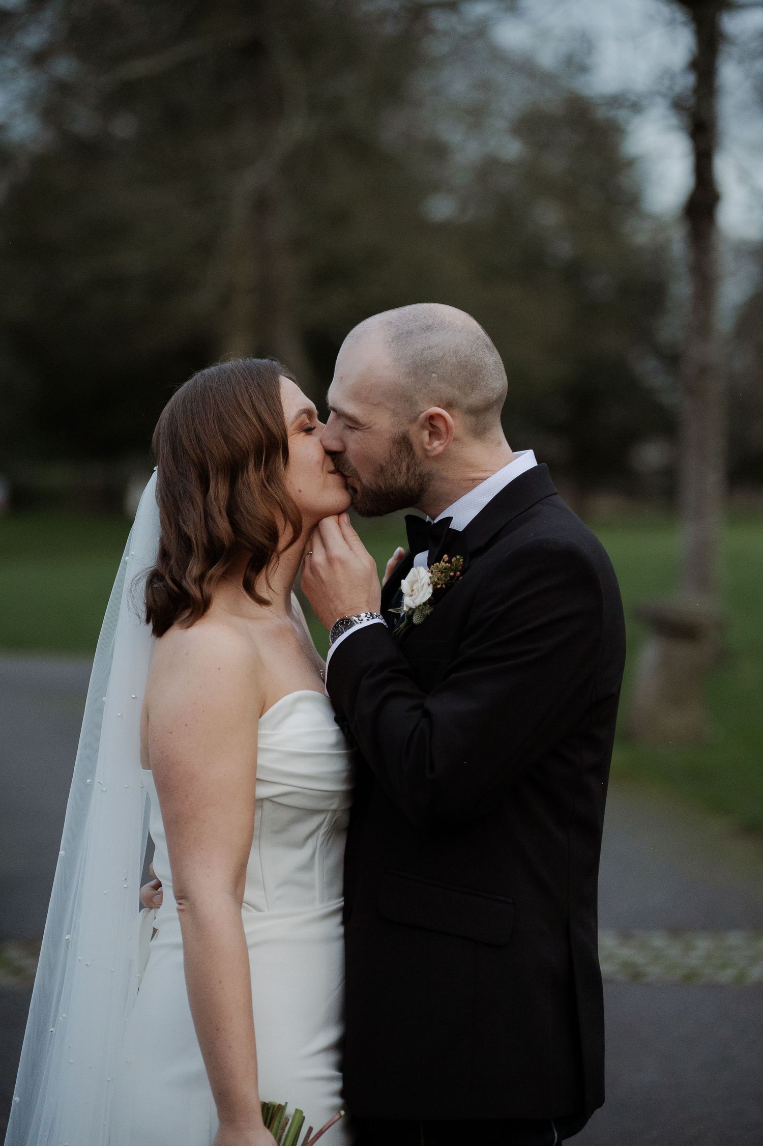 Bride and groom share a romantic kiss outdoors, she in a strapless white gown with veil, he in a black tuxedo.