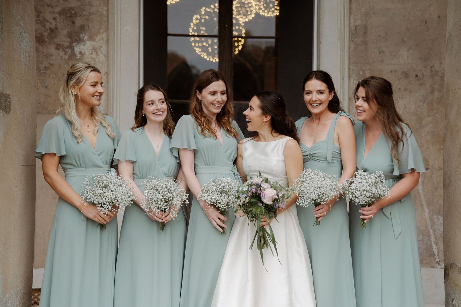 Bride and five bridesmaids in sage green dresses holding bouquets pose together at an elegant wedding venue.
