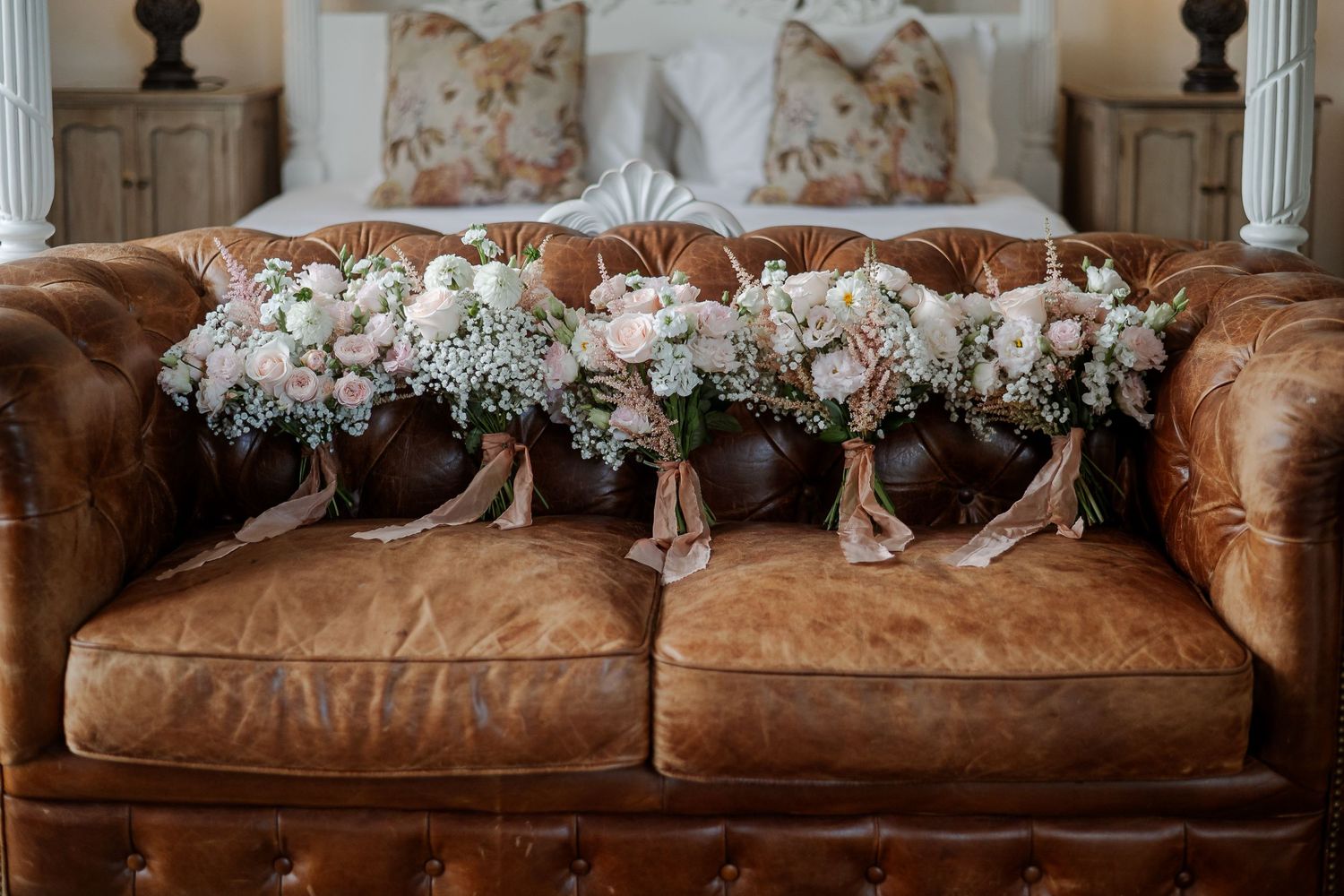 Four romantic blush and white wedding bouquets with ribbon ties resting on a tufted brown leather sofa.