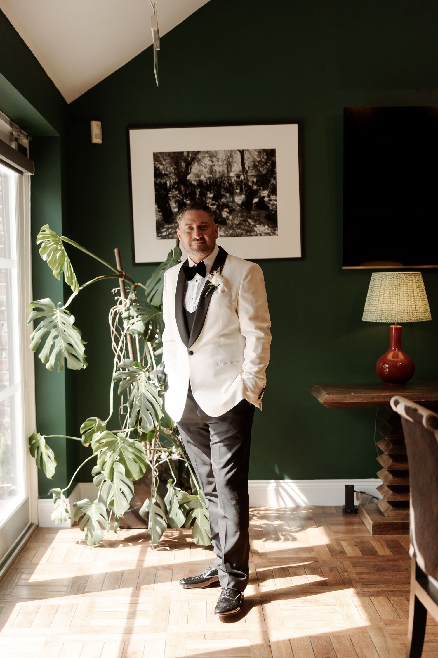 Groom in white tuxedo jacket and black bow tie posing in a room with dark green walls and tropical plants.