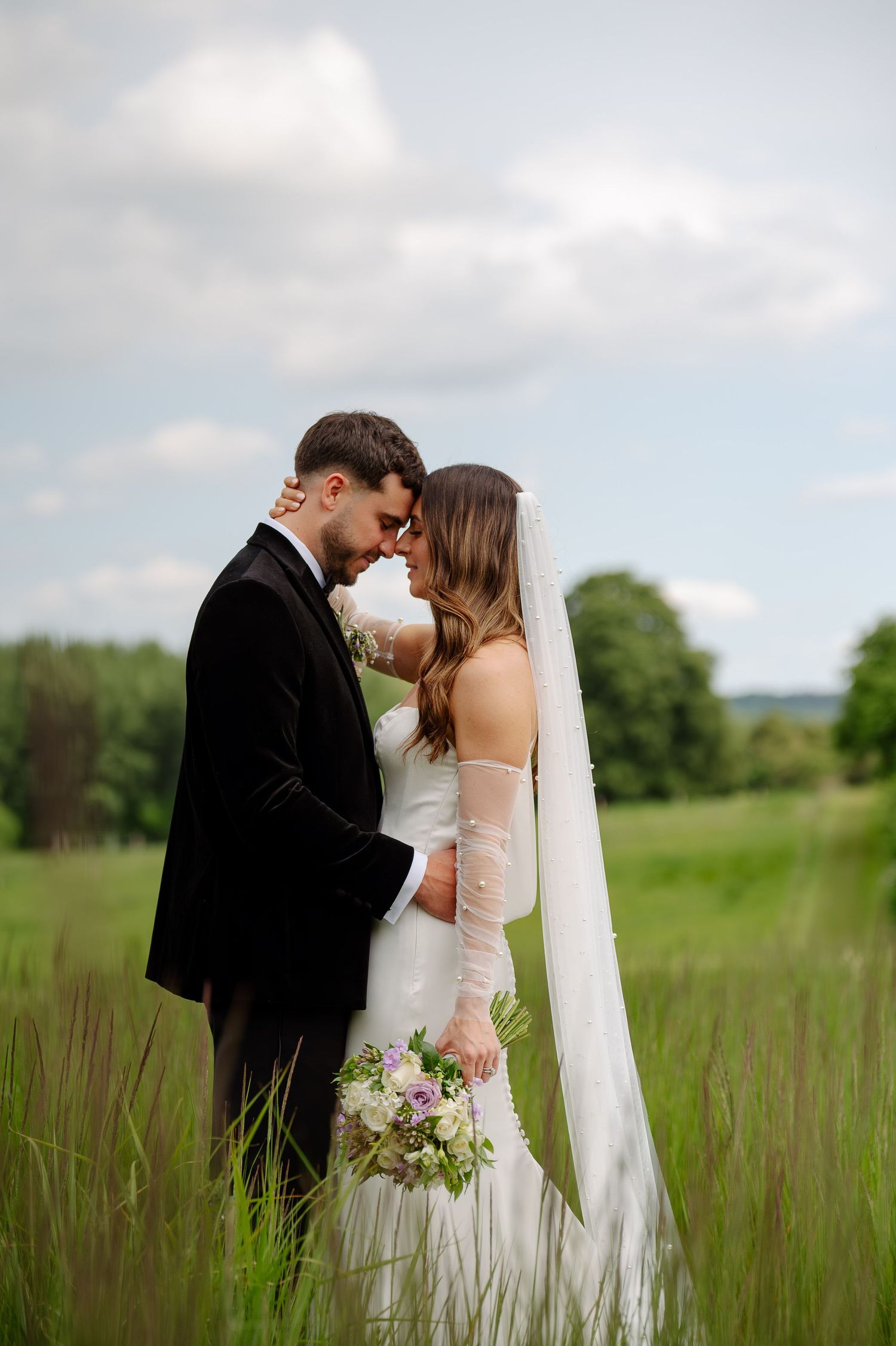 Newlyweds embrace in a serene meadow with tall grass and cloudy skies as backdrop.