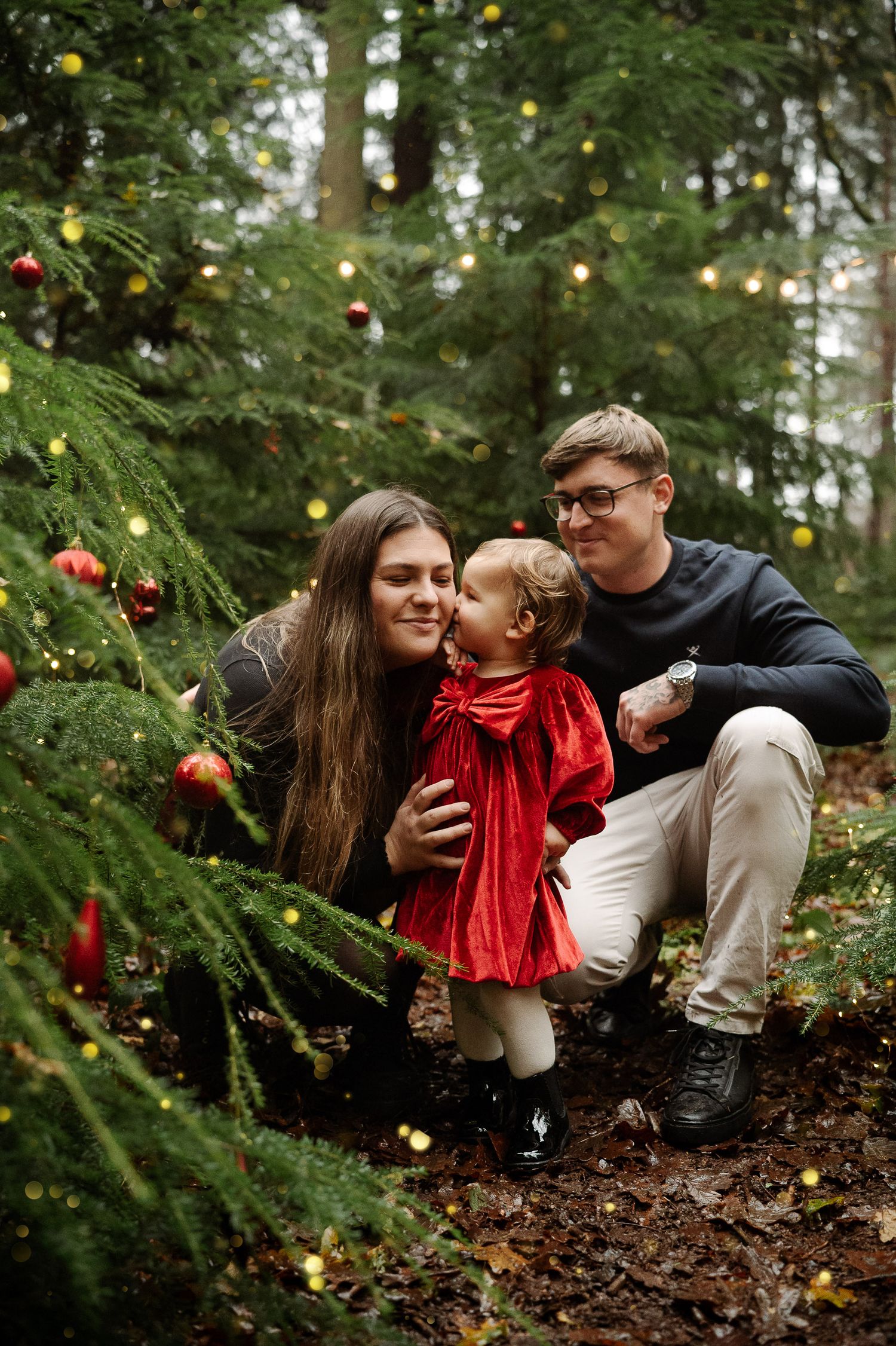 Family with toddler in red dress posing by decorated Christmas tree in forest with holiday ornaments.